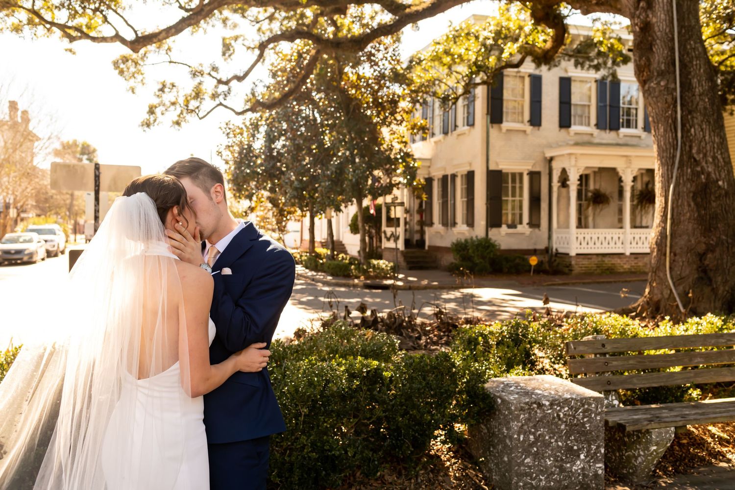 A romantic newlywed couple embraces in a golden sunlit garden with historic mansion buildings in the background.