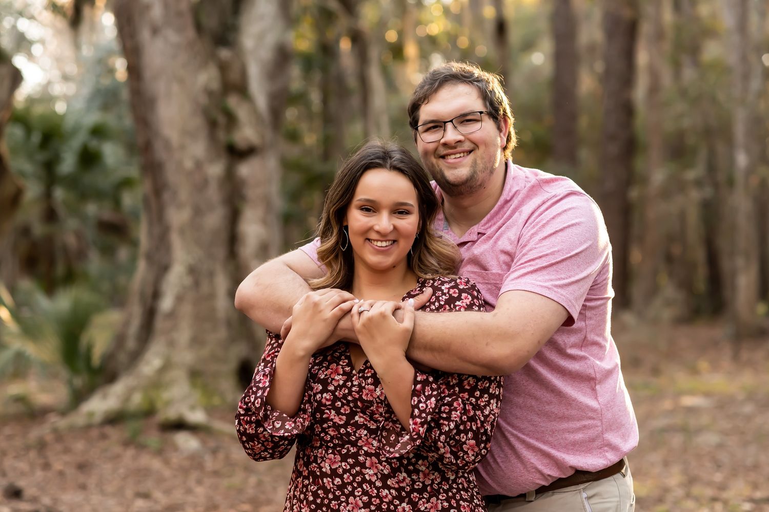 A couple poses together making a heart shape with their hands while standing in a wooded forest setting.