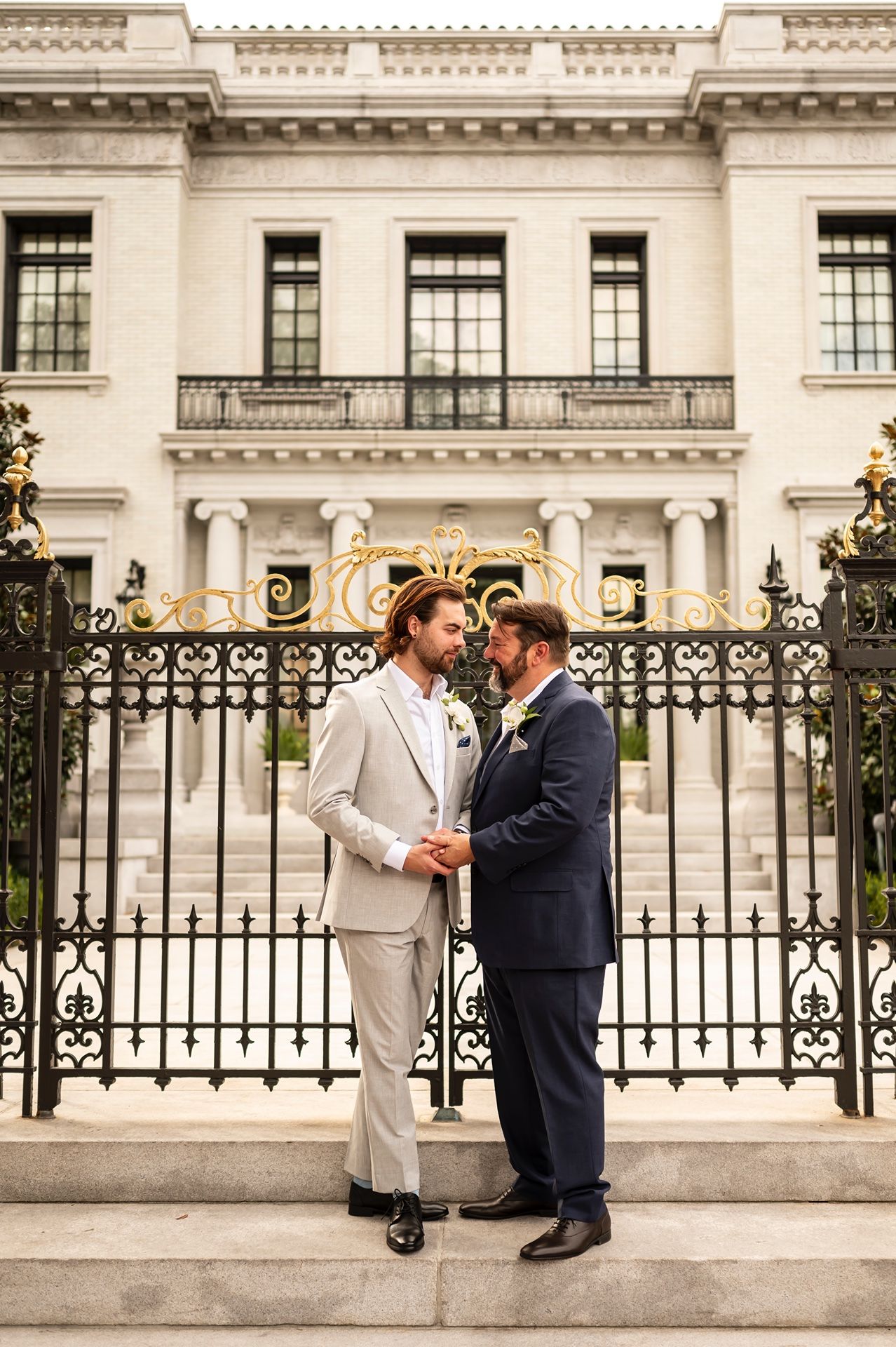 A couple embraces on marble steps in front of a grand white building with ornate iron gates.