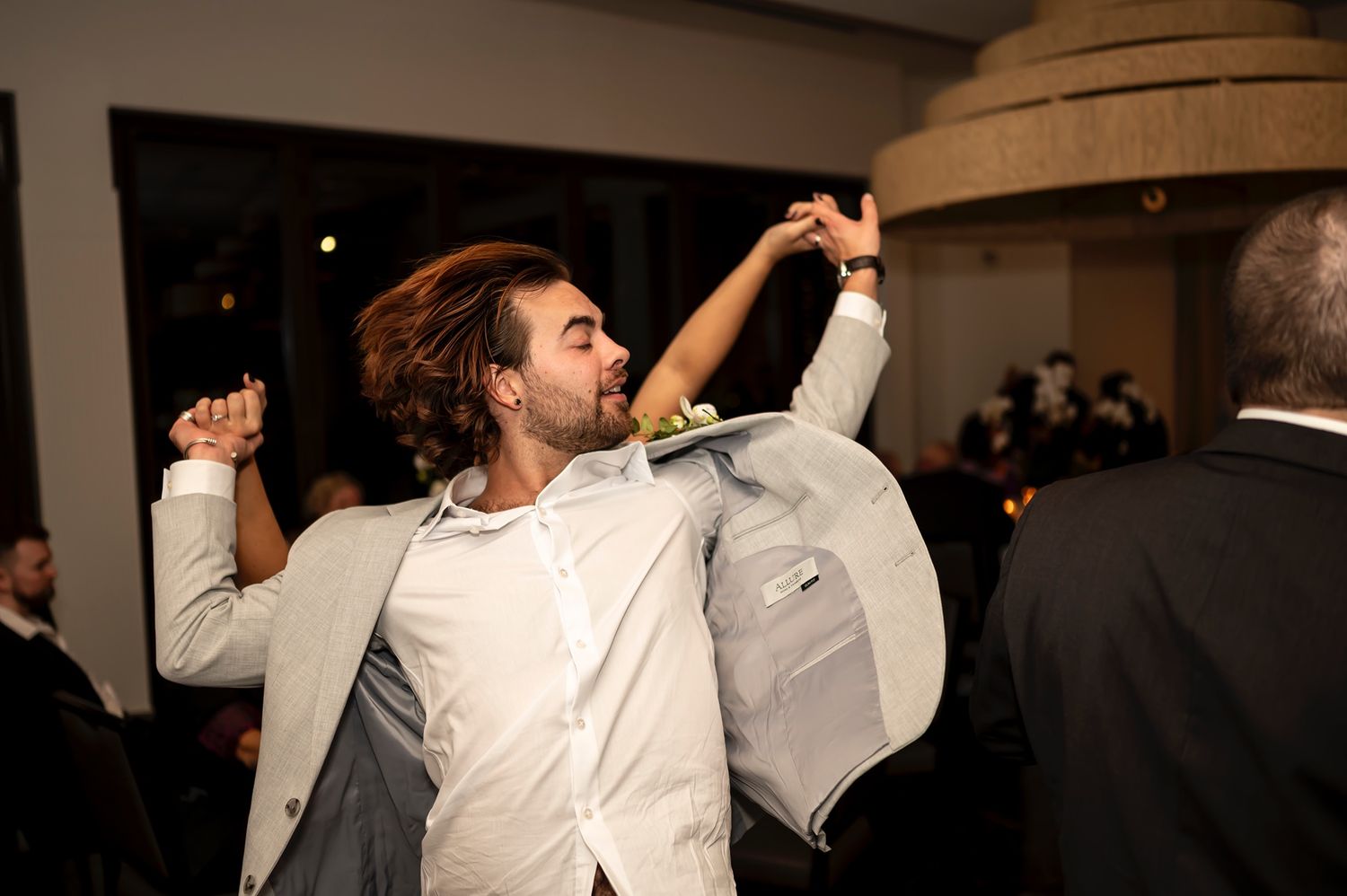 A person in a white shirt and light-colored blazer dances energetically at a dimly lit event.