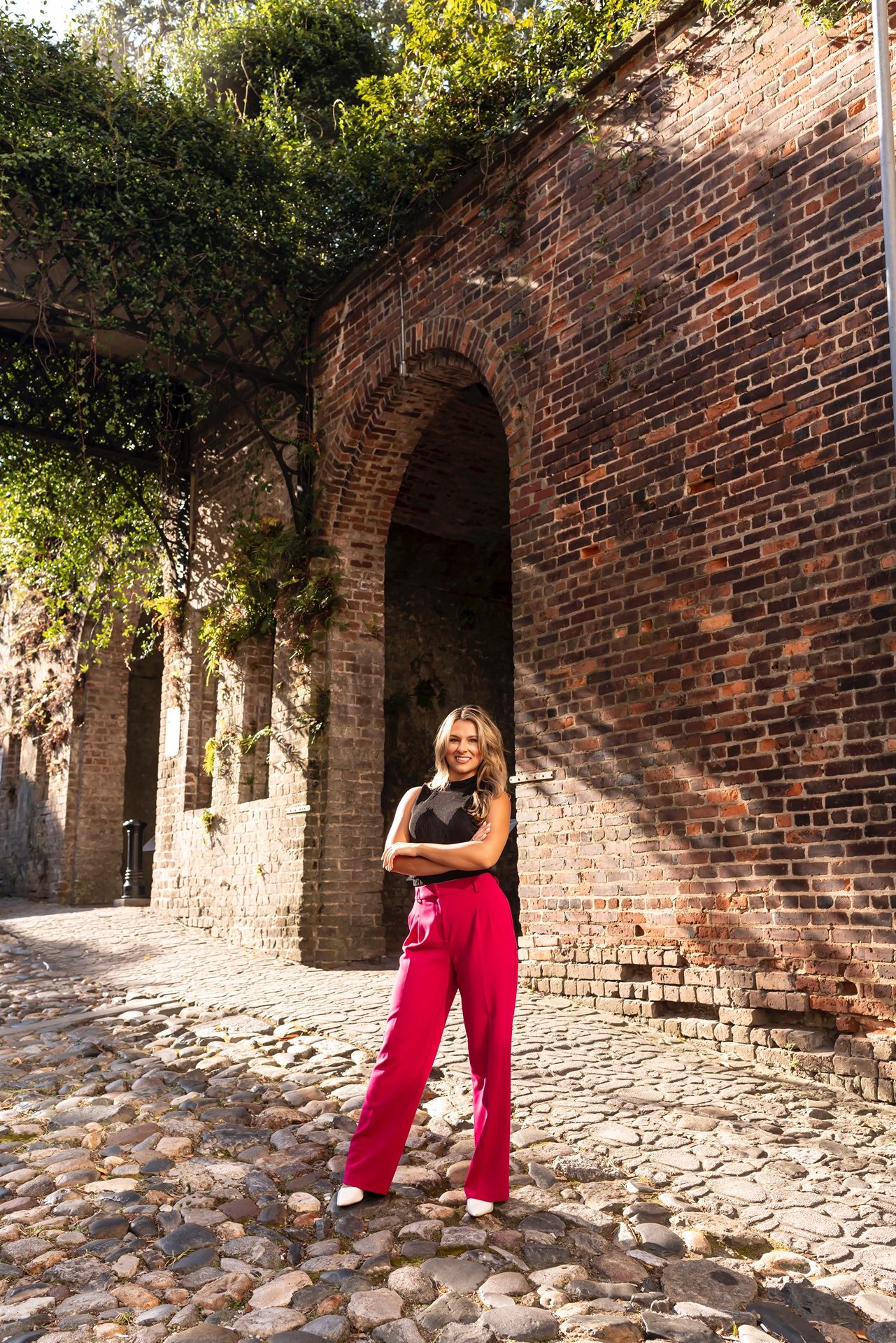 Person in red pants poses against rustic brick archway with cobblestone path and greenery in historic setting.