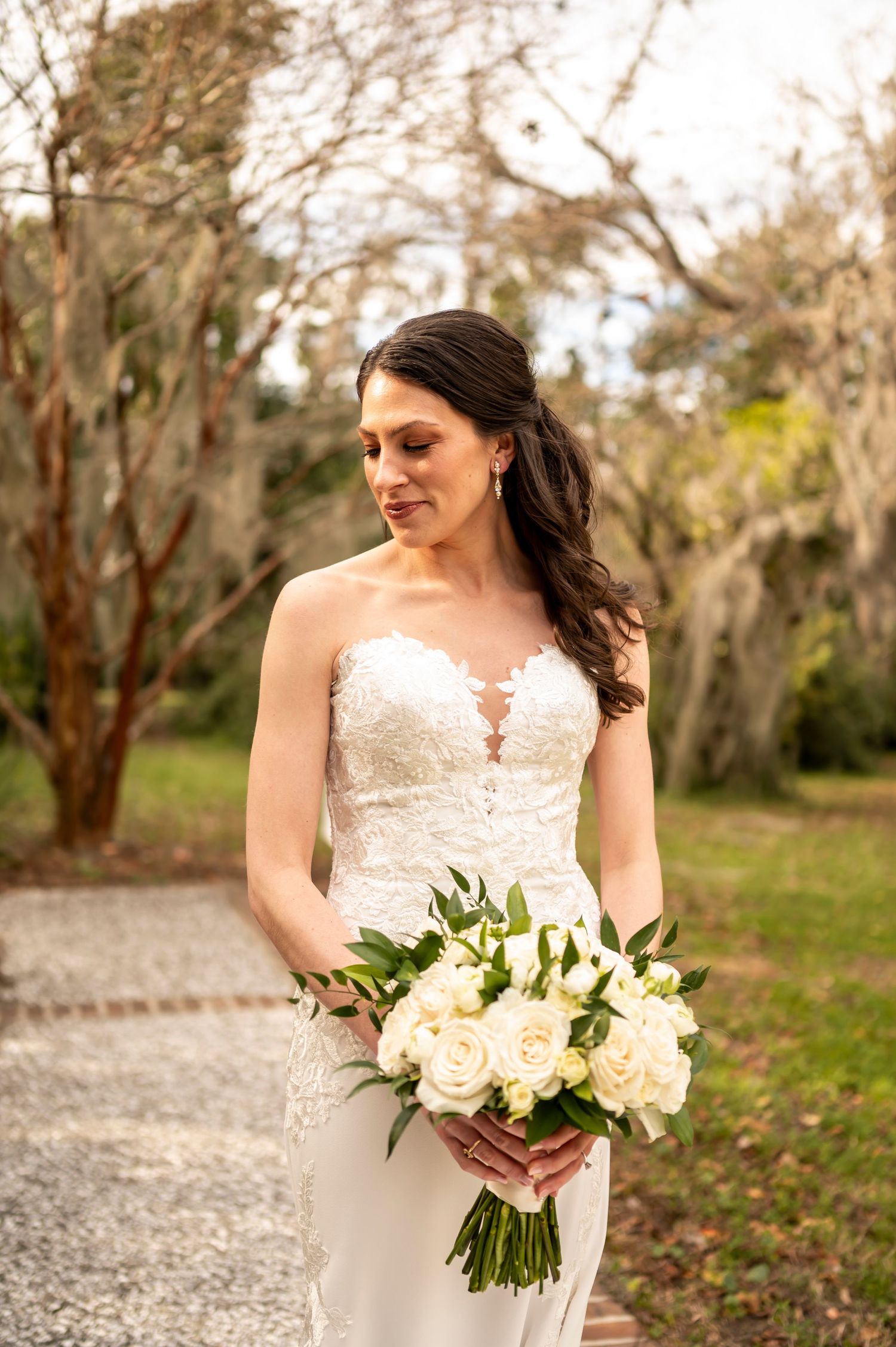 A bride in a strapless lace wedding gown holds an elegant white and cream floral bouquet while standing on a gravel path.