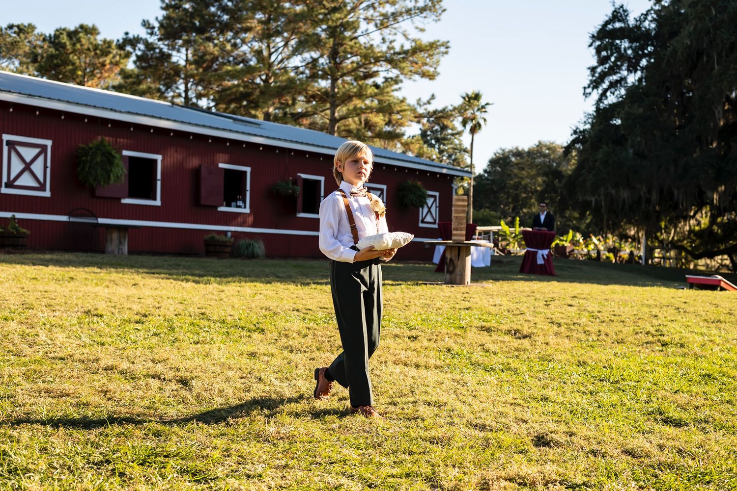 Young boy in formal attire stands on grassy lawn in front of red barn building during golden hour.