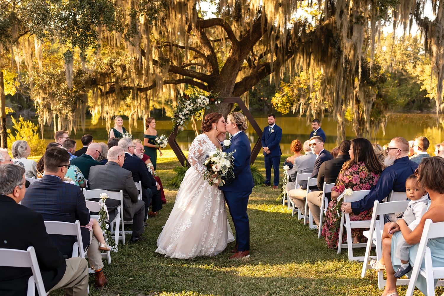 Outdoor wedding ceremony taking place under moss-draped oak trees with white folding chairs arranged on the grass at sunset.