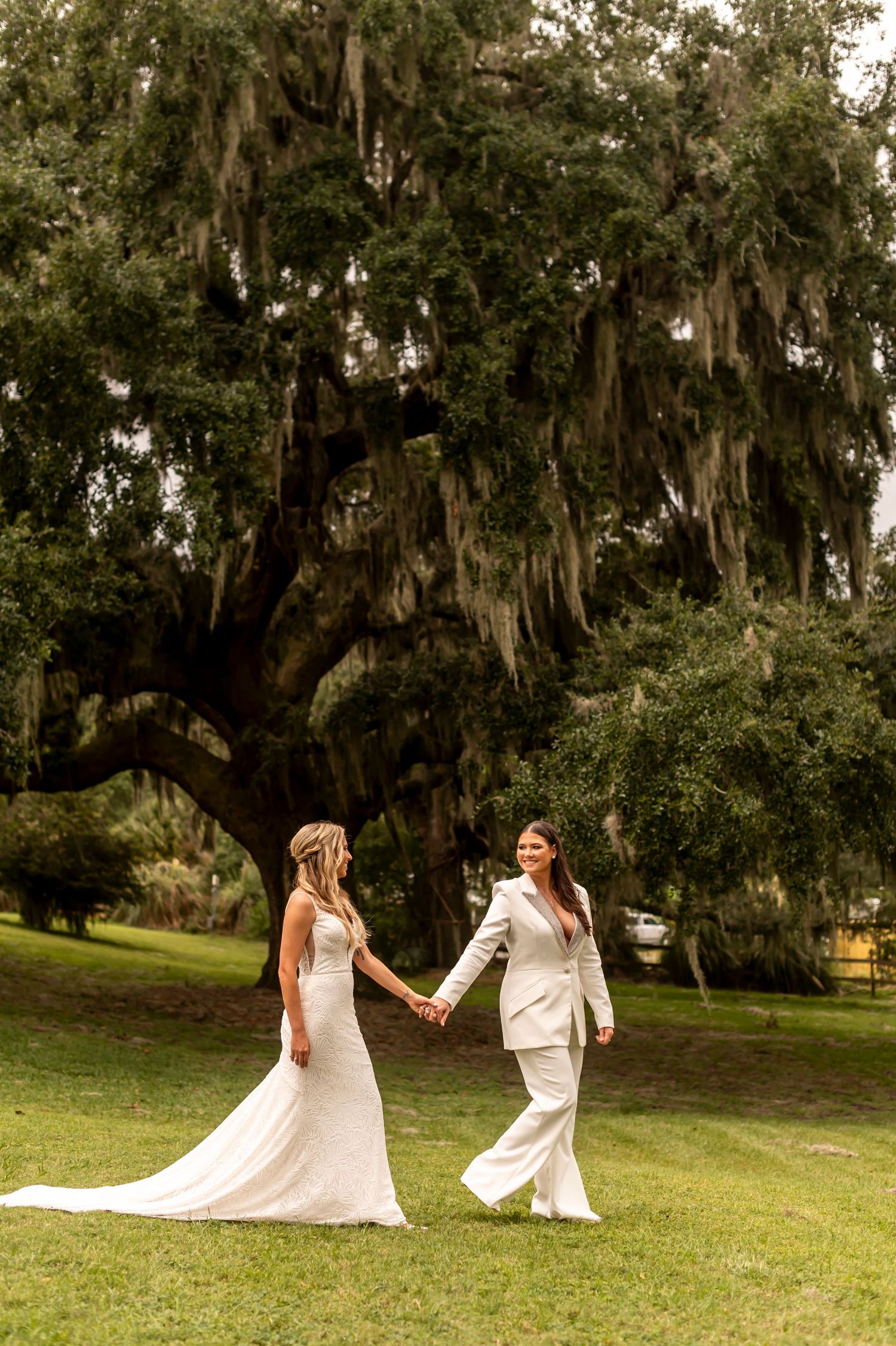 A same-sex wedding couple holds hands while walking on grass beneath a majestic oak tree with Spanish moss.
