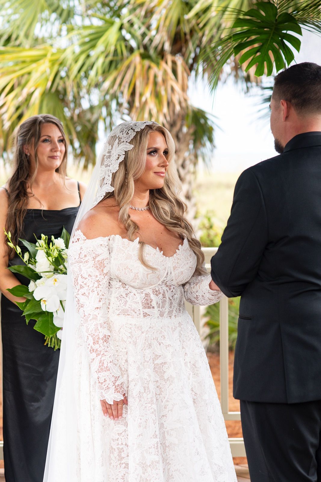 A bride in a long-sleeved lace wedding gown stands with guests at an outdoor venue with palm trees.