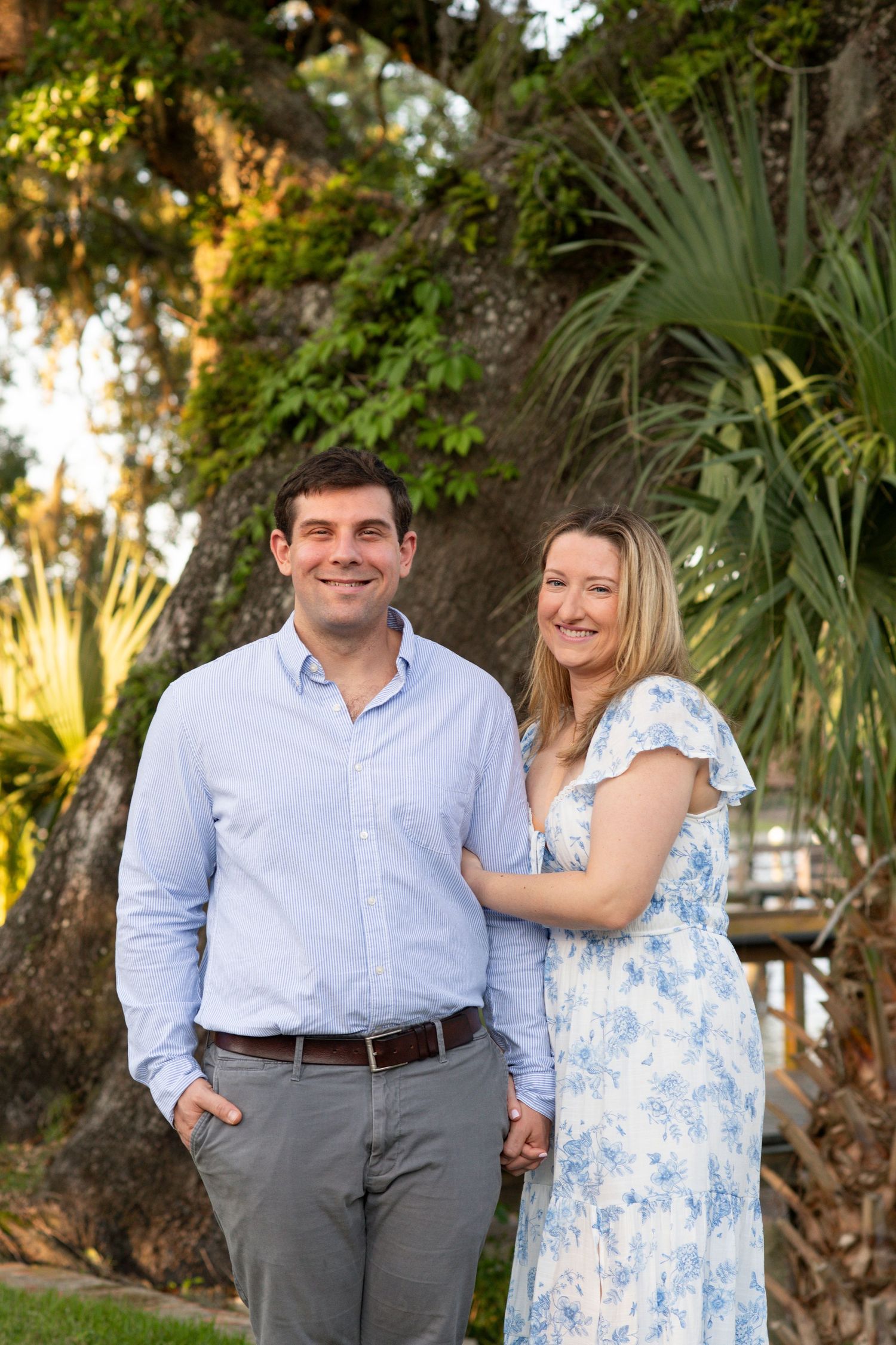A couple in coordinating blue and white outfits poses together in a lush garden setting during golden hour.