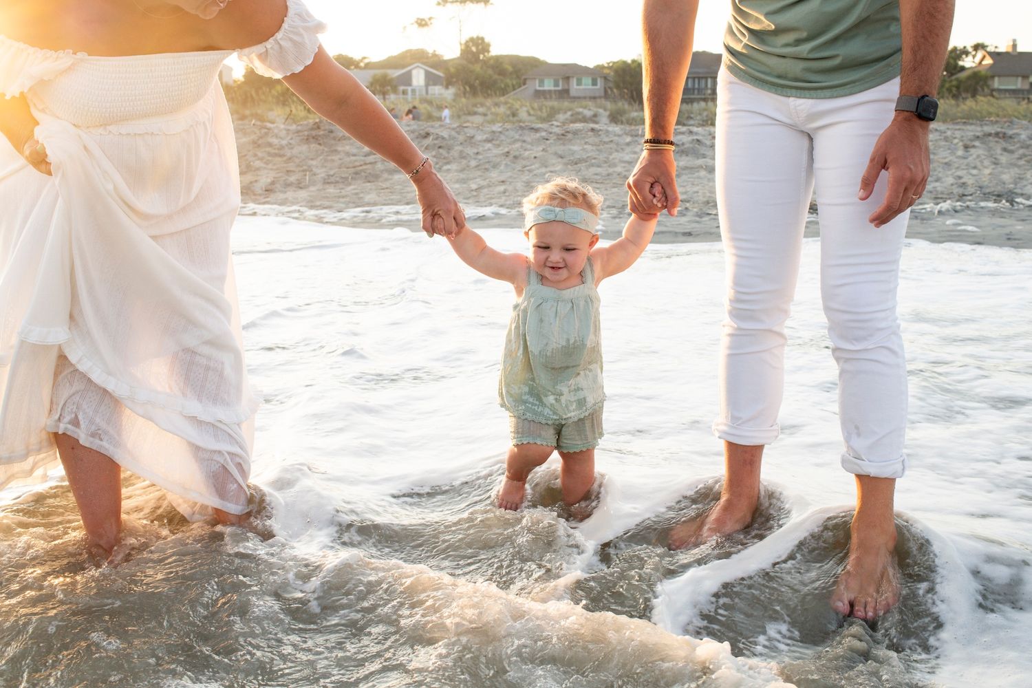 Two adults holding a baby's hands while walking in ocean waves on a sunny beach.
