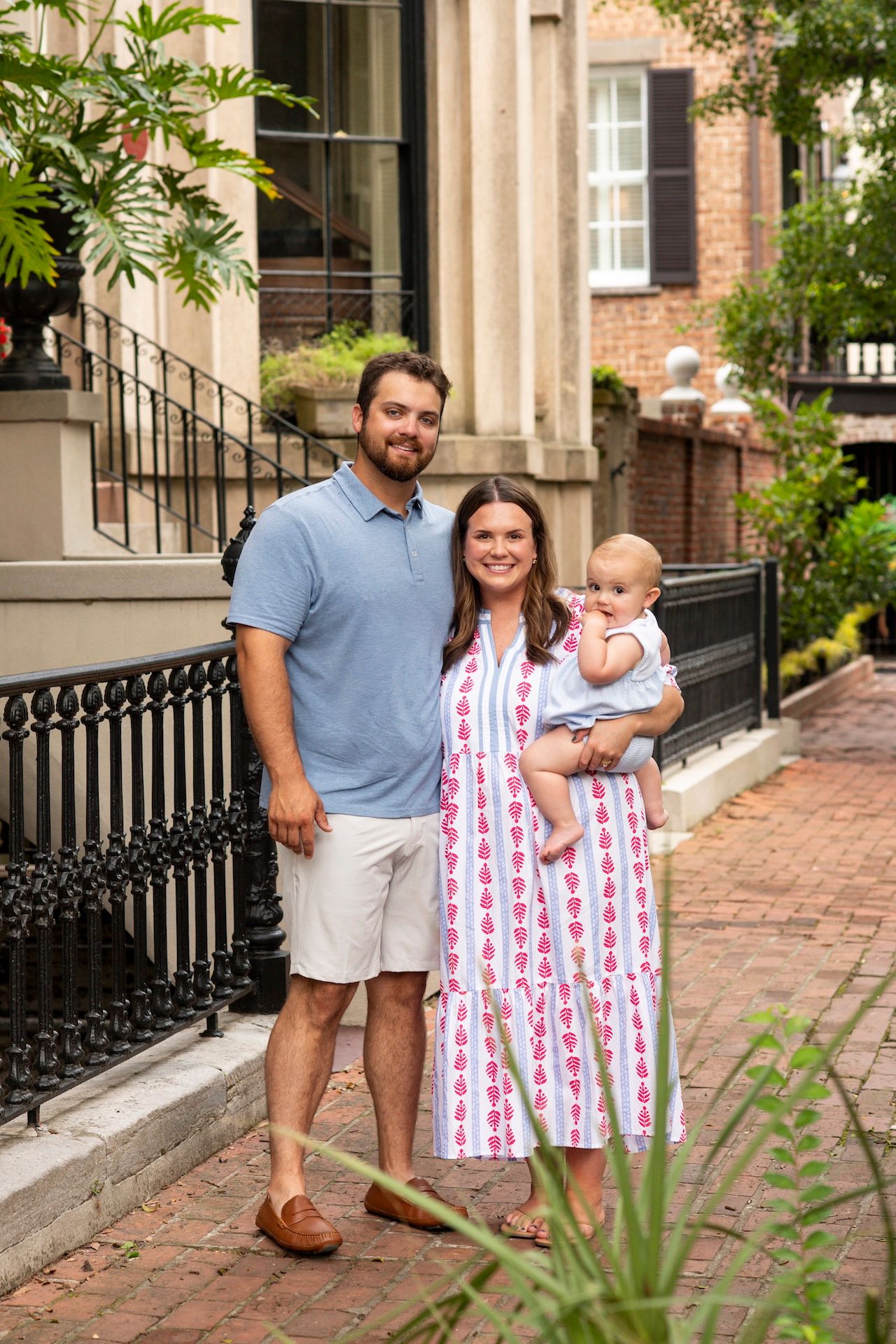 A young family of three poses on a brick sidewalk, with man in blue polo, woman in pink dress holding baby.