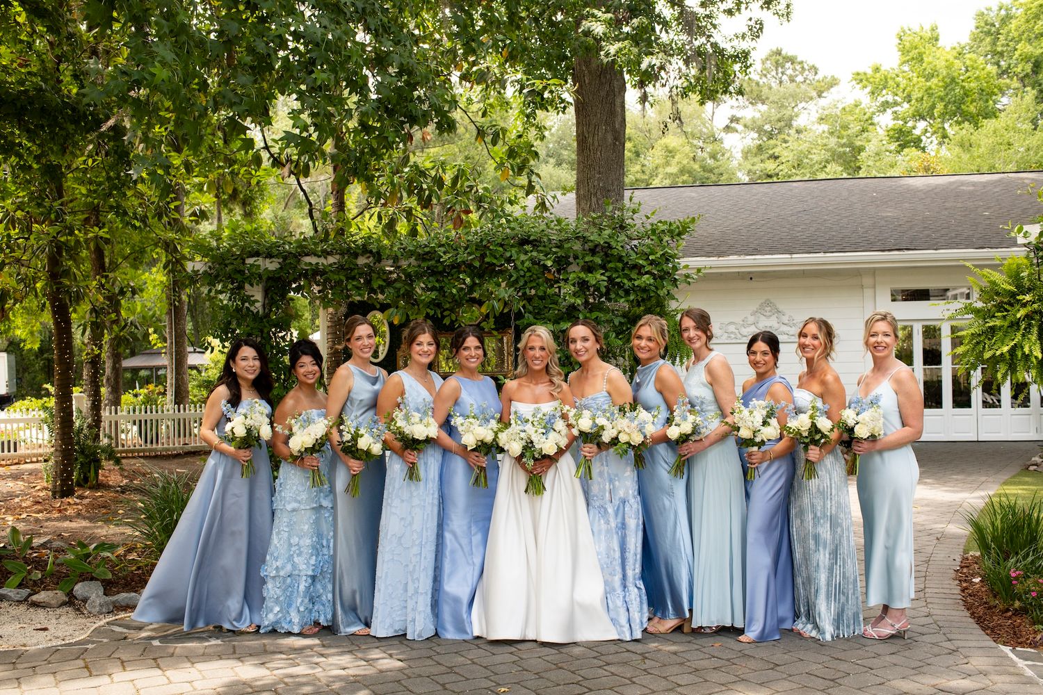 Bride in white gown poses with bridesmaids in blue dresses holding floral bouquets outdoors at a wedding venue.