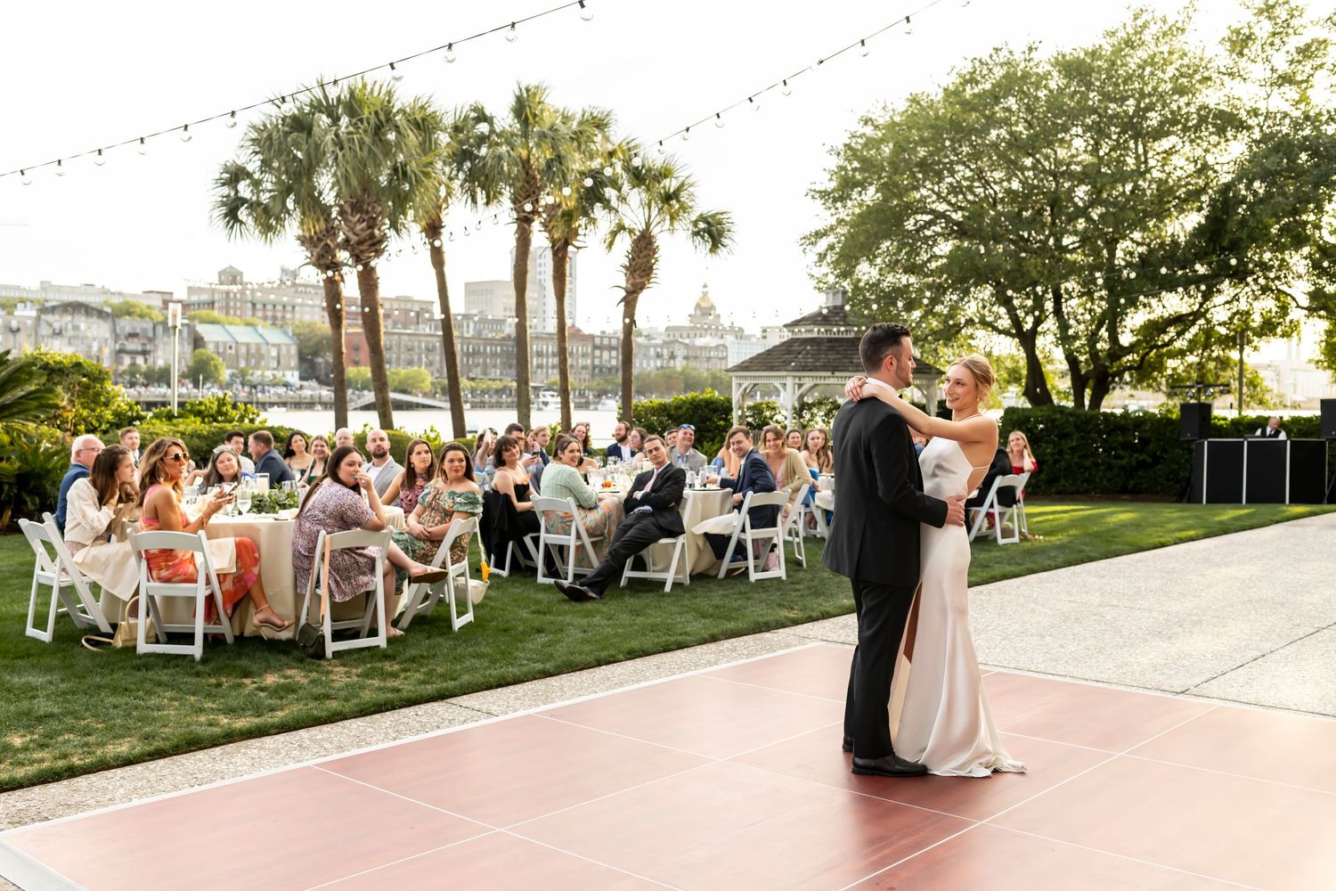 Outdoor wedding reception on a lawn with string lights and palm trees at dusk with downtown Savannah, Georgia in the background.
