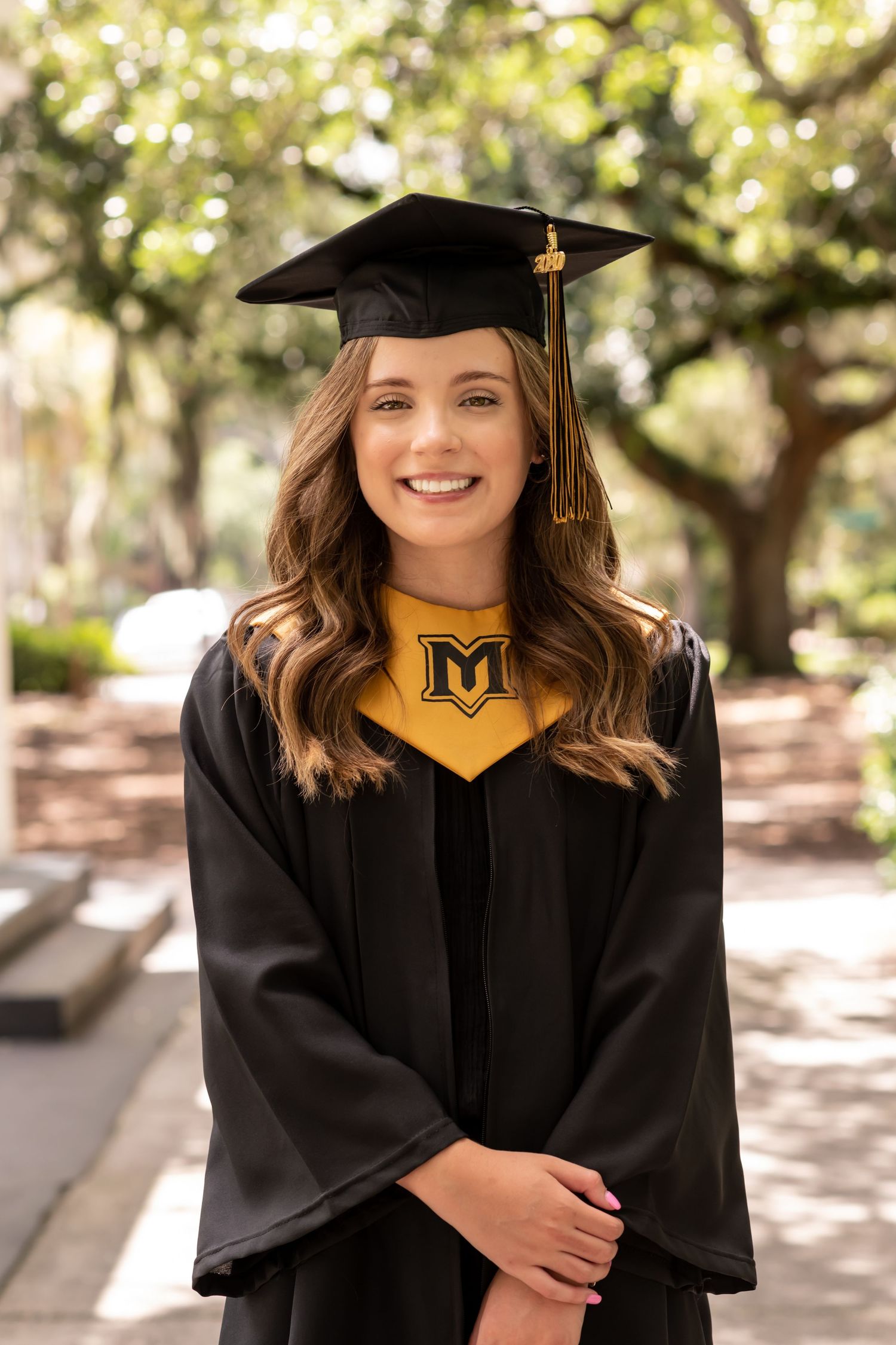 Graduate in black cap and gown with gold M emblem smiling outdoors on campus.
