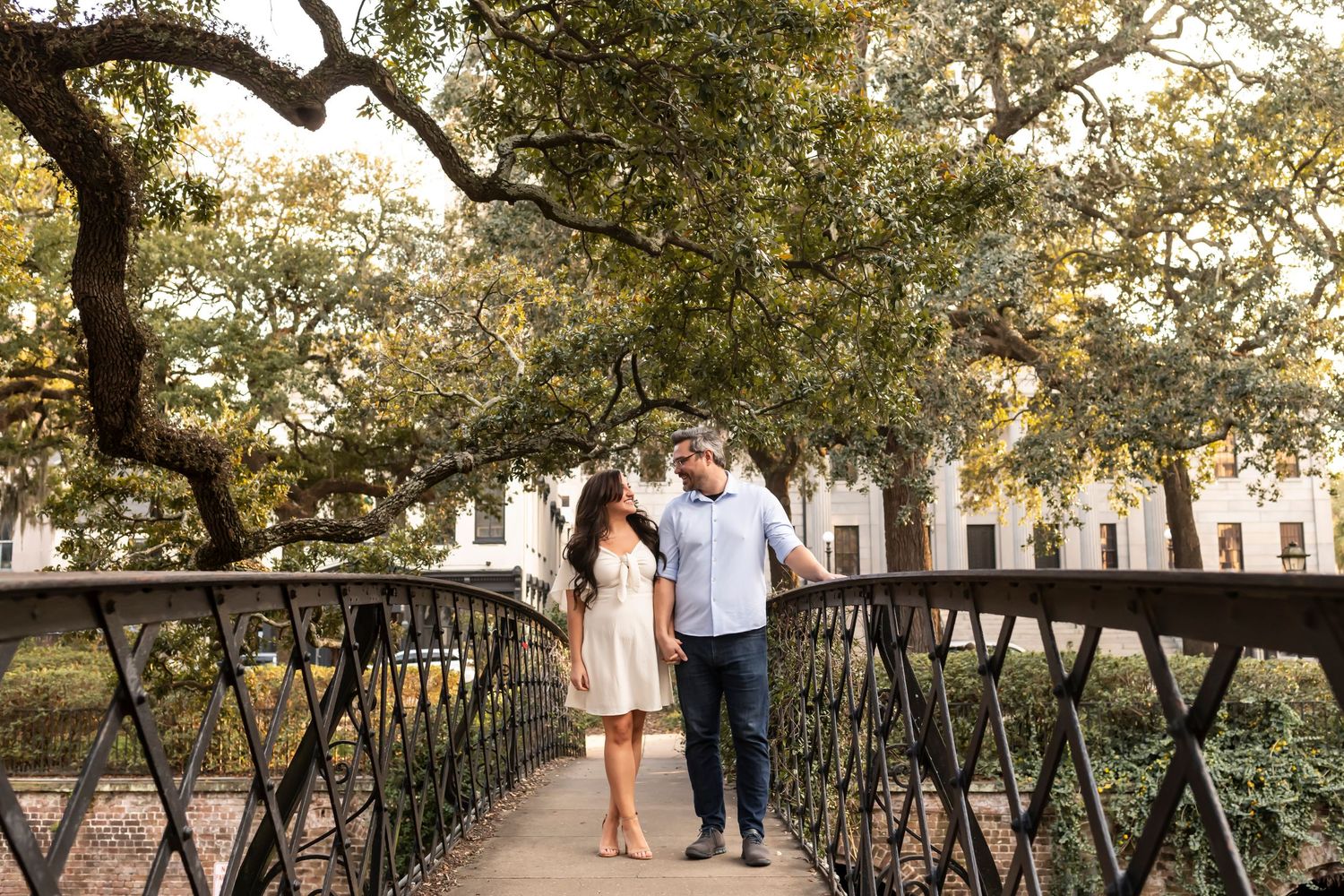 A couple stands together on an ornate iron bridge surrounded by lush greenery and trees on Factor's Walk in Savannah, Georgia.