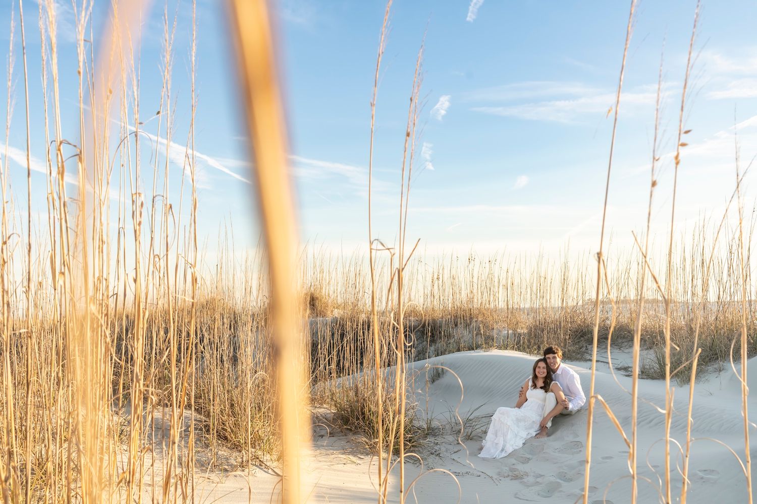Couple sitting romantically in sandy beach dunes surrounded by golden sea oats under a blue sky.
