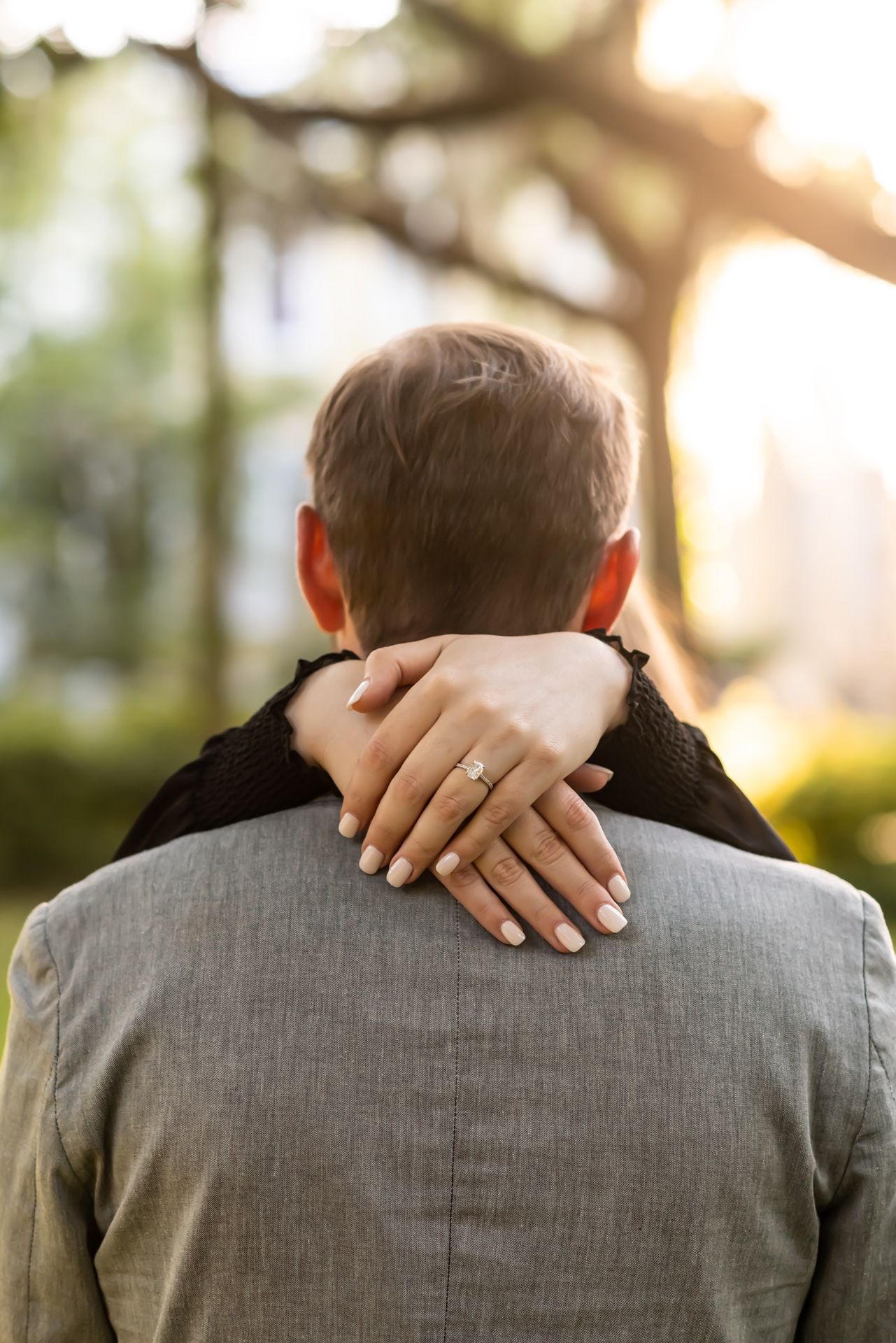 A person's hands gently rest on someone's shoulders from behind, wearing formal gray suit attire in soft sunlight.