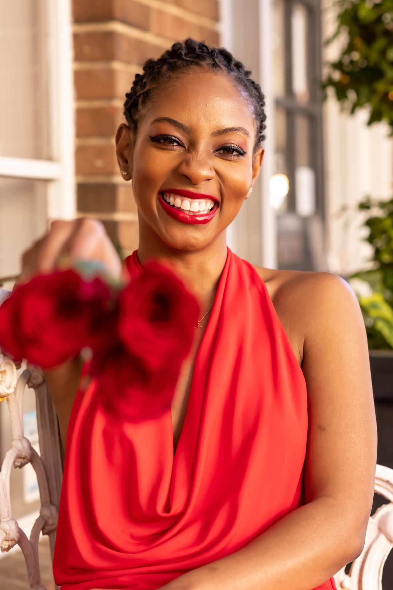 A radiant smile shines in a red sleeveless cowl neck top against a brick exterior background.
