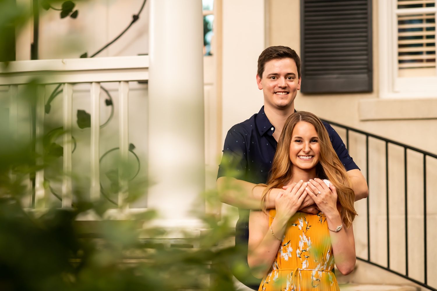 A happy couple embraces on their porch during a sunny engagement photoshoot.