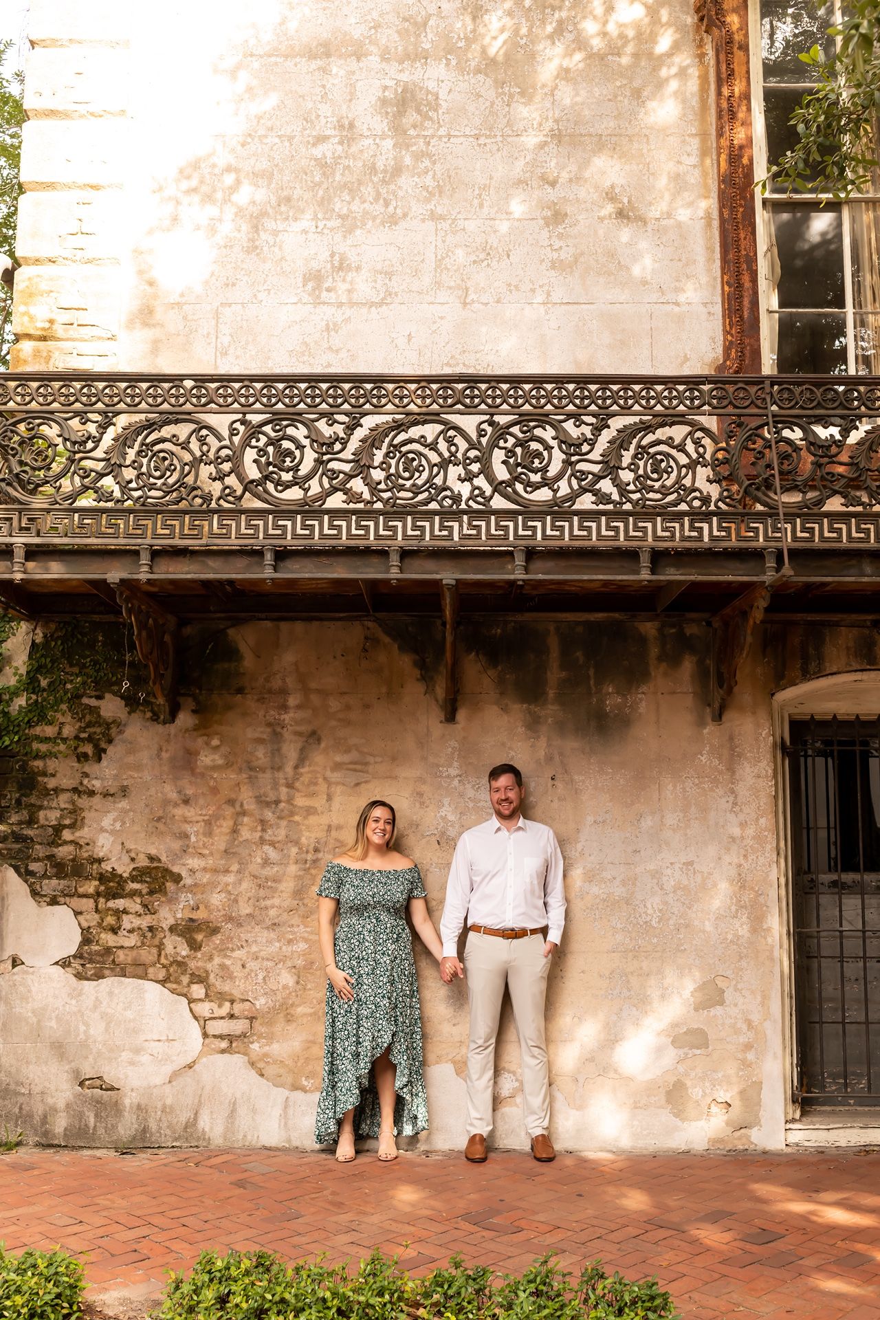 Couple stands beneath ornate wrought iron balcony on historic building with weathered walls and terracotta tiles.