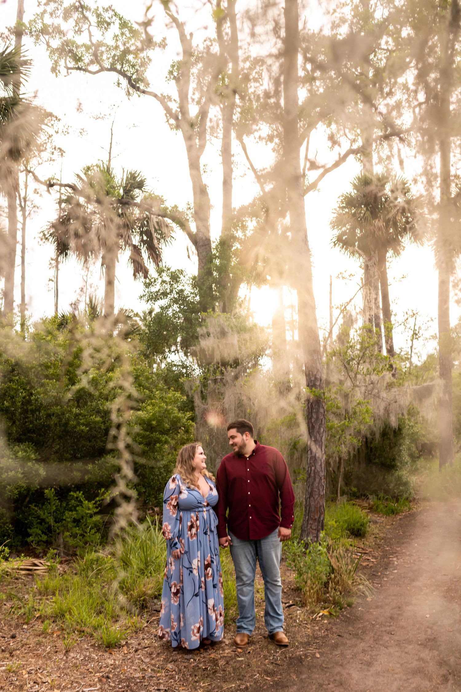 A couple walks together on a rustic dirt path surrounded by palm trees and Spanish moss at sunset.