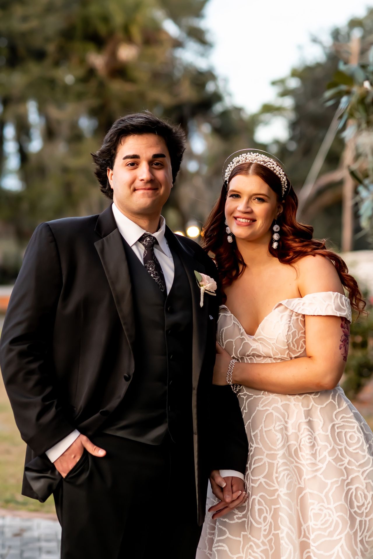 A bride in a white lace dress and groom in black suit pose outdoors in a nature setting during their wedding photoshoot.