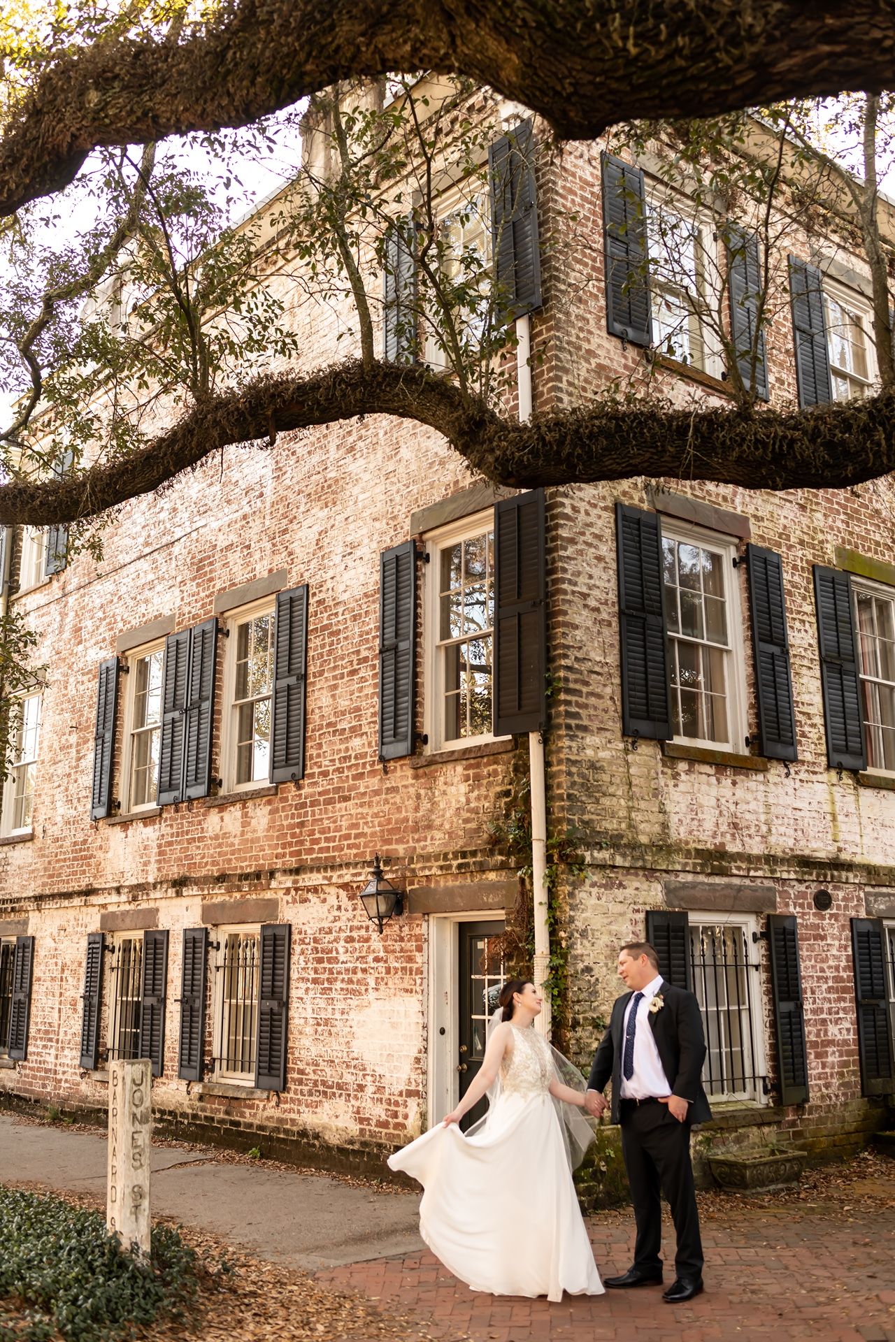 Wedding couple poses in front of historic brick building with large oak tree during sunset photo session.