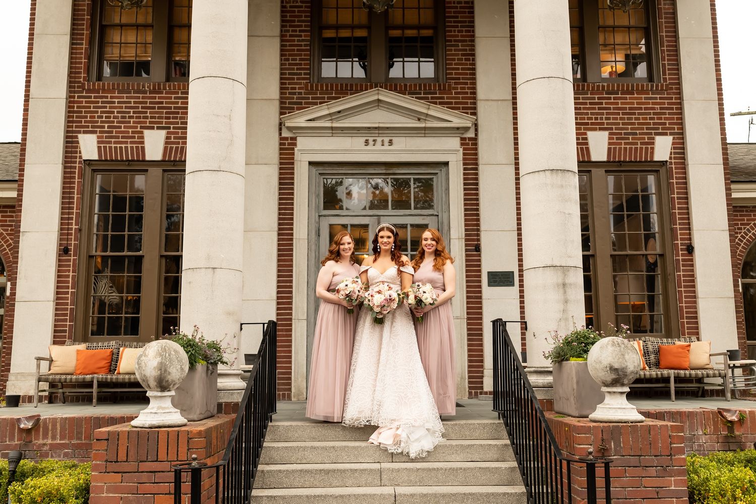Bridal party in matching pink dresses poses on elegant brick building steps with white columns and decorative urns.