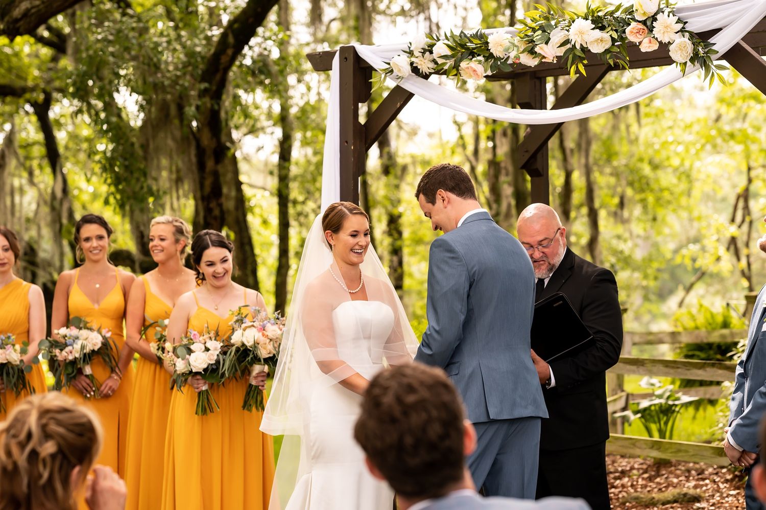 An outdoor wedding ceremony under trees with bridesmaids in yellow dresses watching the couple and officiant.