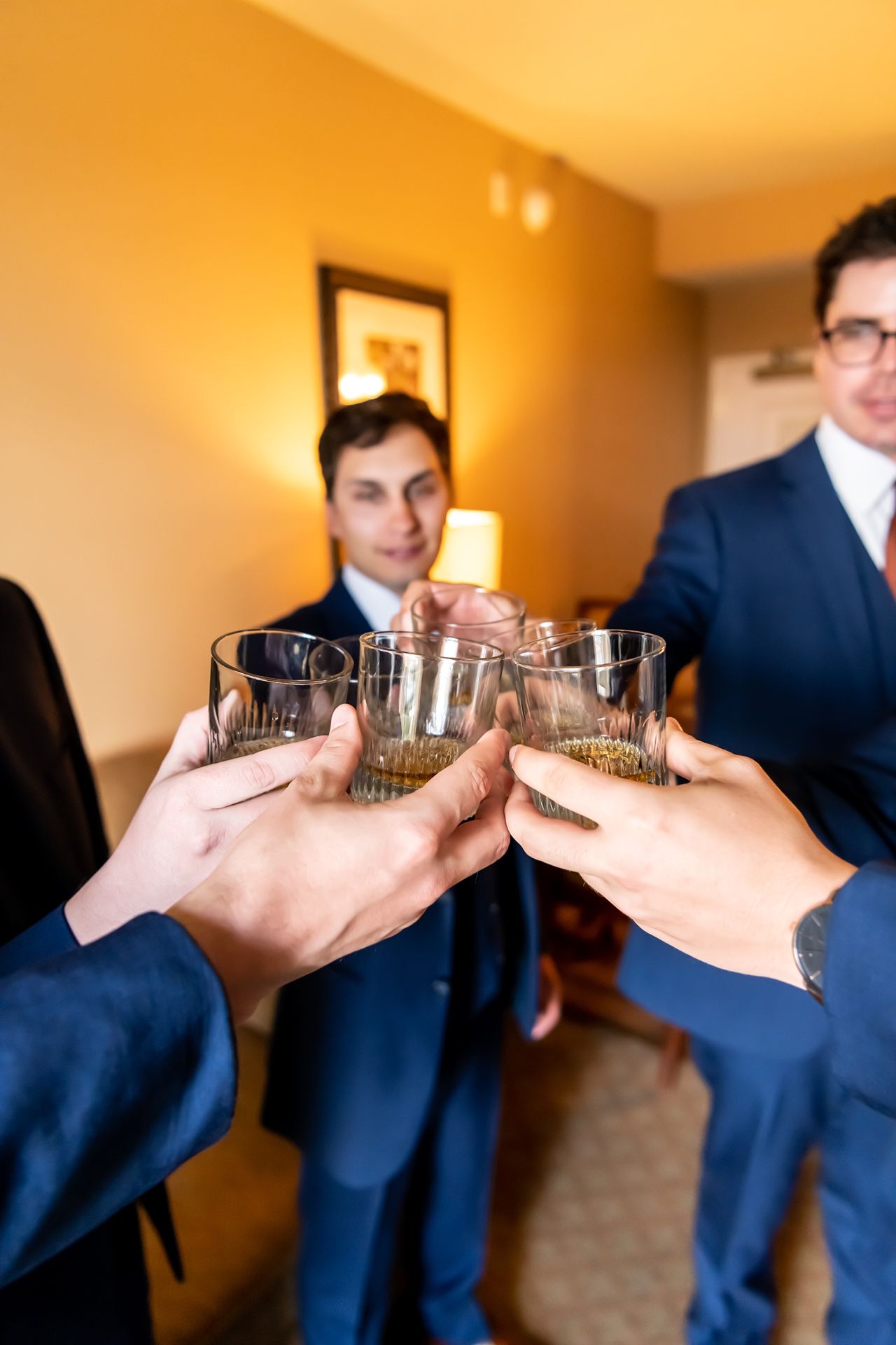 People in navy blue suits celebrate with a toast of whiskey glasses in a warmly lit room.