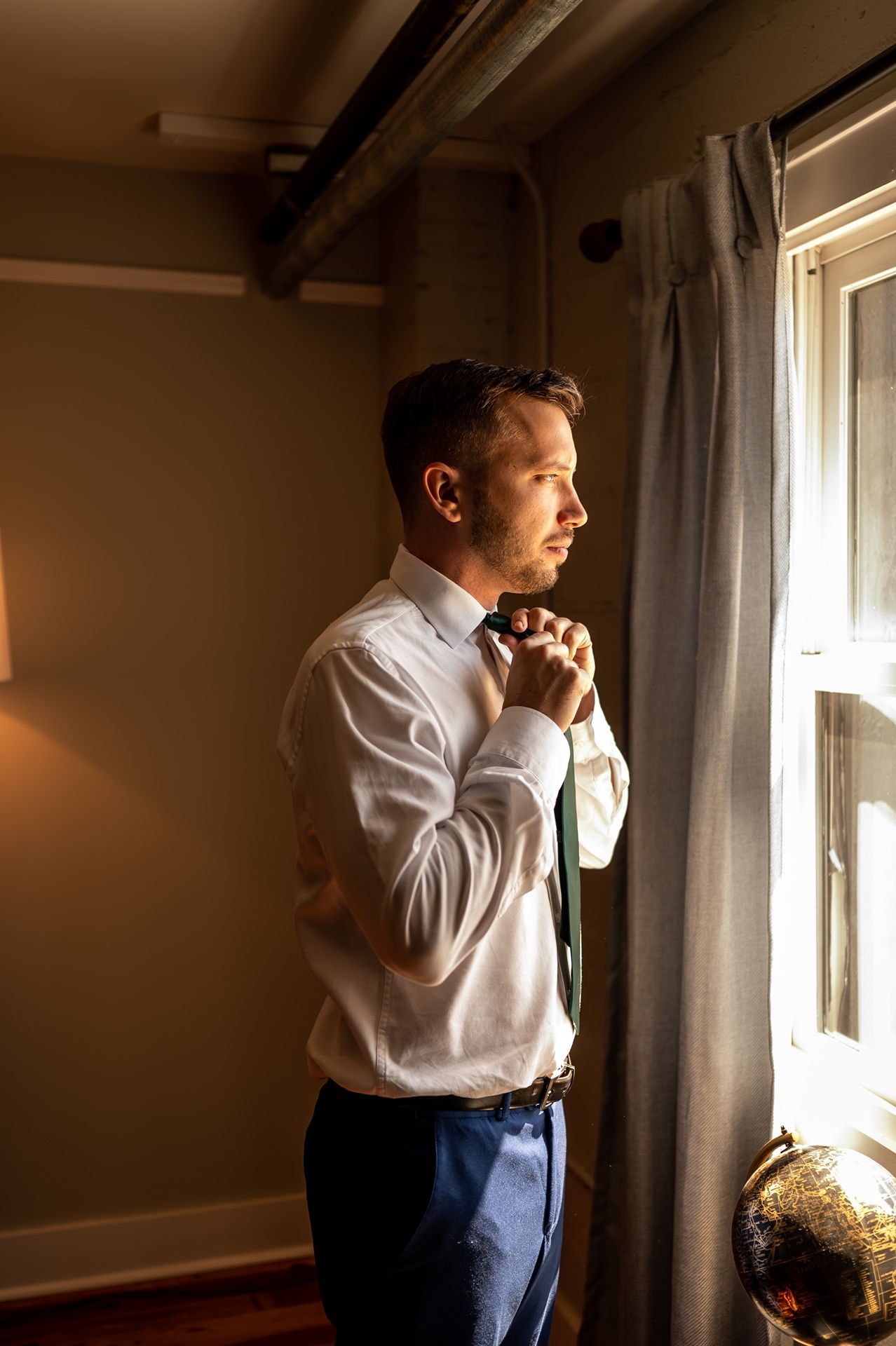 A person in a white dress shirt and dark pants stands near a window gazing at sunlight streaming in.