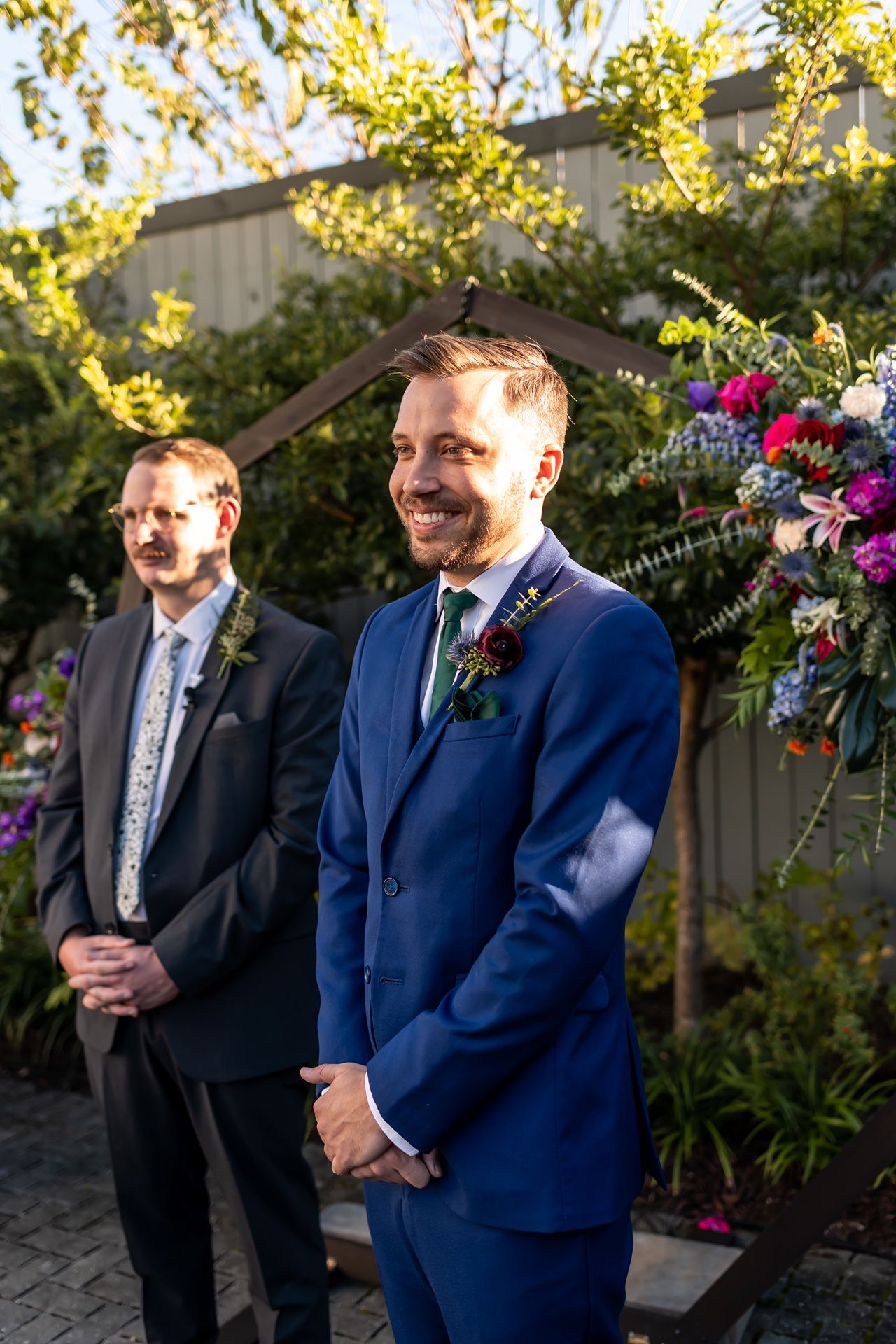 Two groomsmen stand together in formal attire outdoors with vibrant floral arrangements during an autumn wedding.