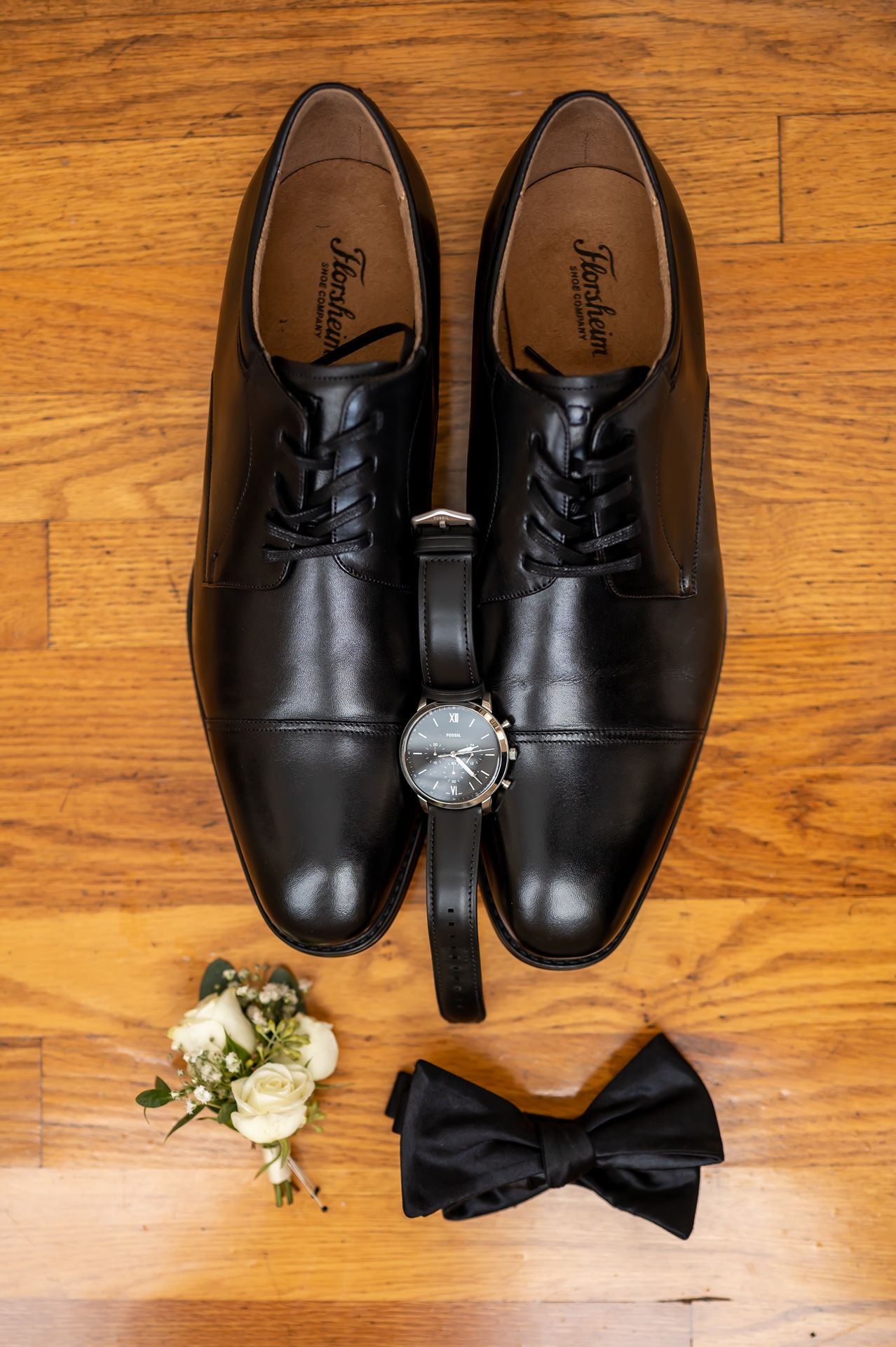 Black formal dress shoes with a watch, bowtie and white boutonniere arranged on wooden flooring.