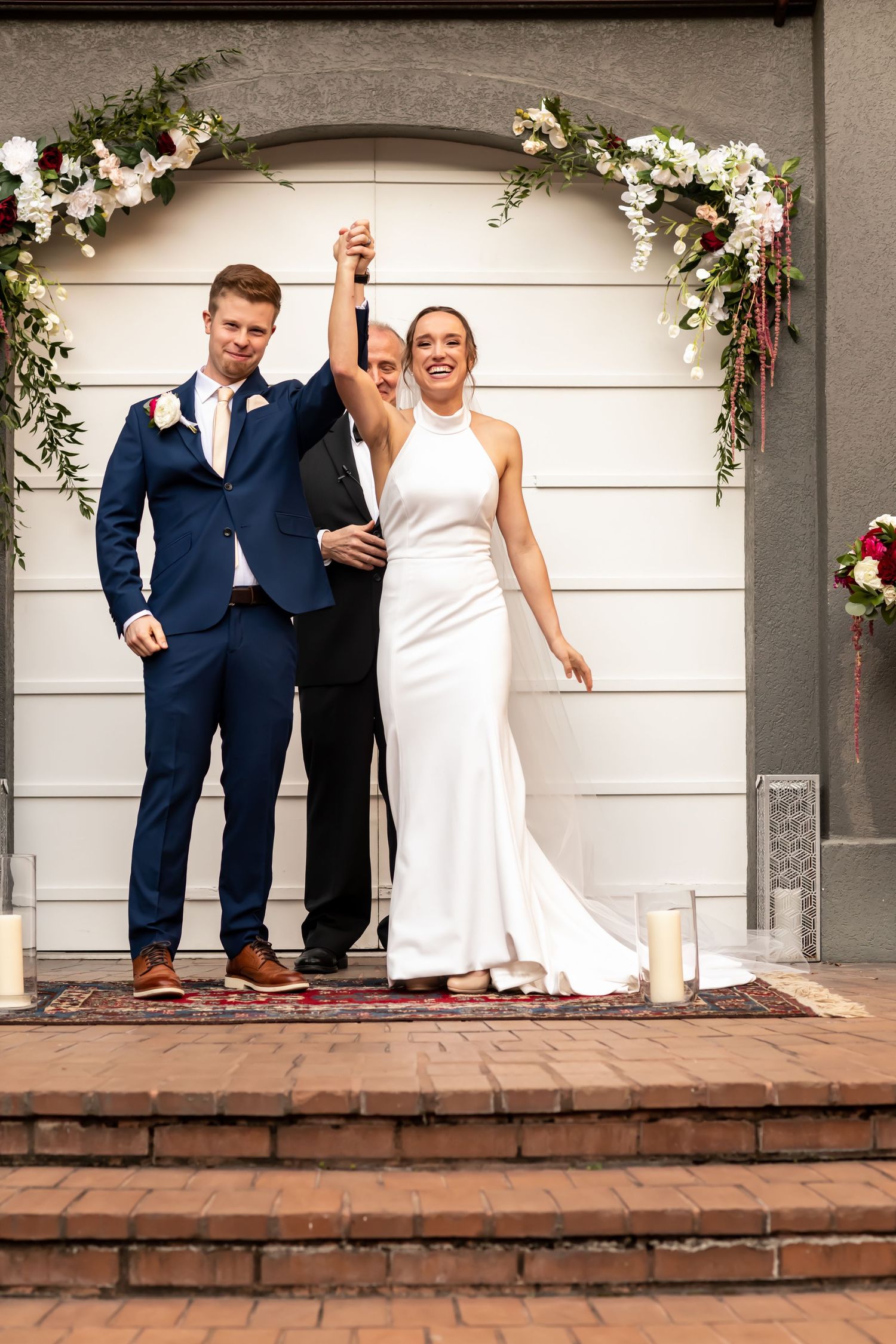 Two people stand joyfully under a floral wedding arch on brick steps in formal attire.
