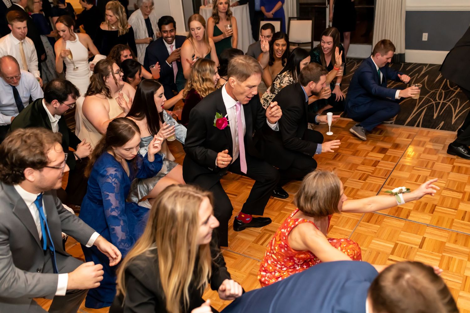 A crowd of formally dressed wedding guests dancing and enjoying themselves on a parquet dance floor during a reception.