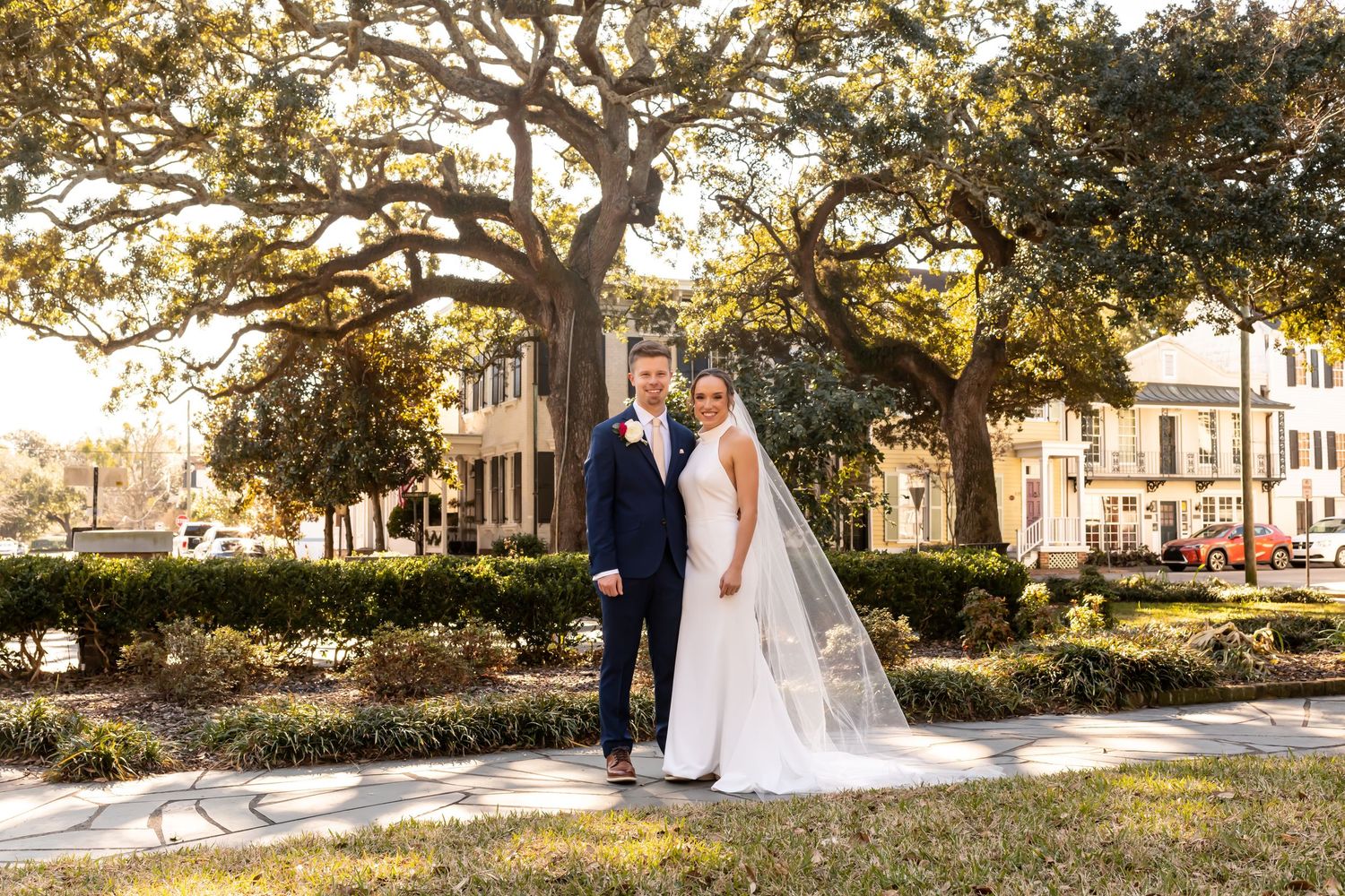A couple in formal wedding attire walk through a serene garden path under sunlit oak trees during golden hour.