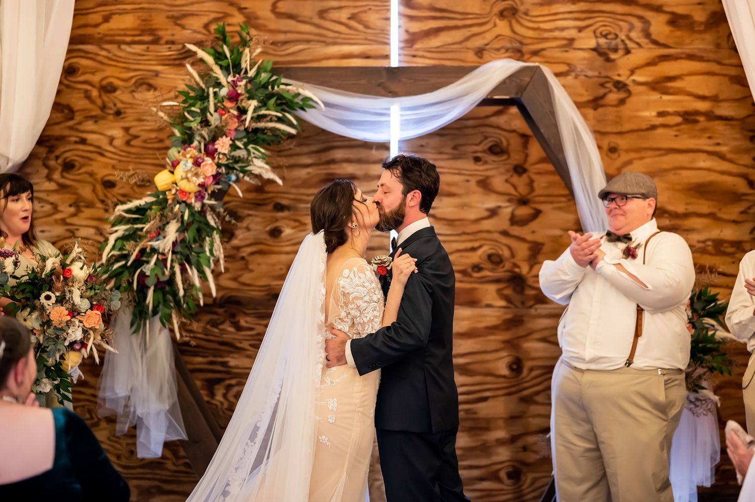 Rustic barn wedding ceremony features elegant floral arch with white draping and peach roses against wooden wall panels.
