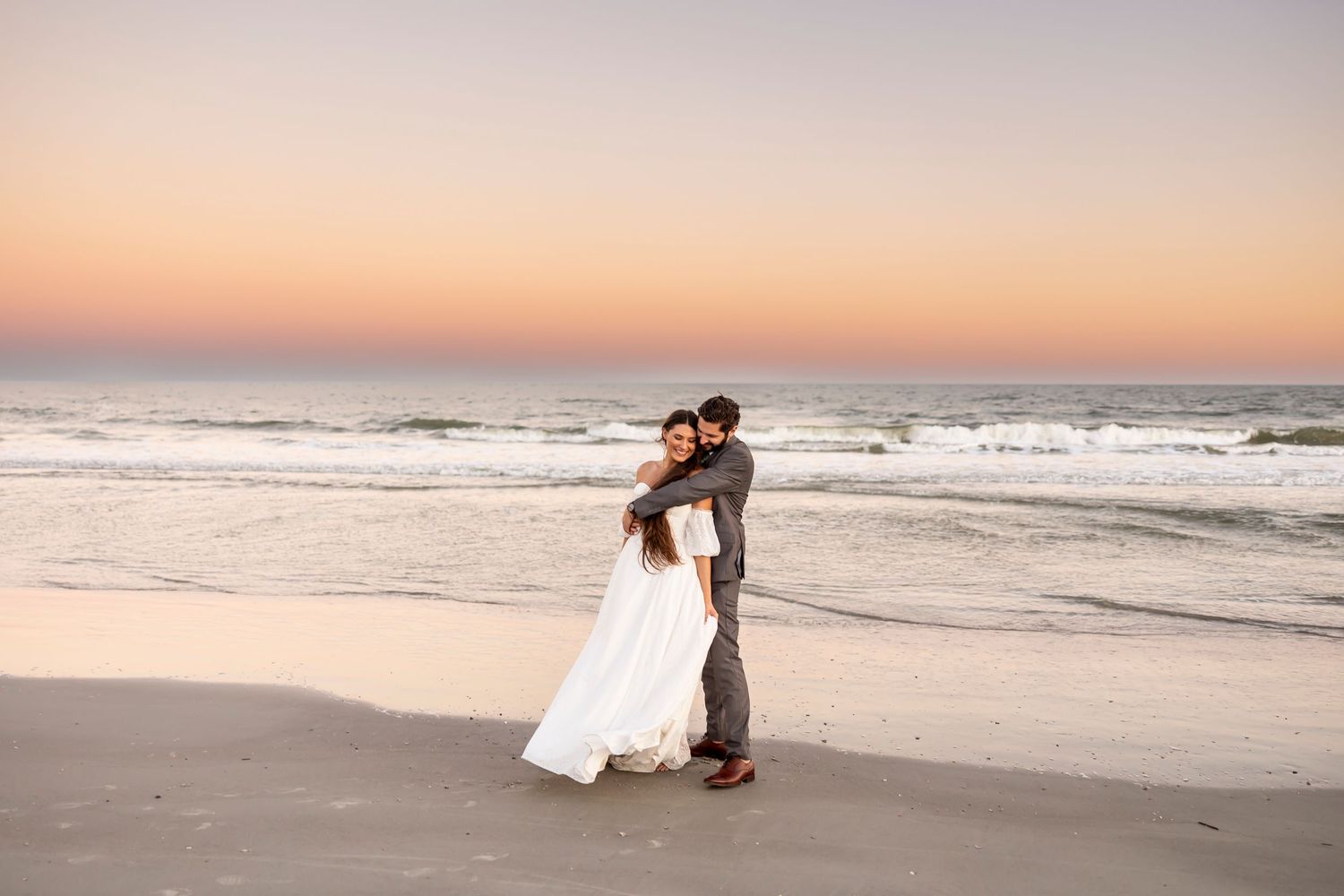 Bride and Groom embrace on a sandy beach on Hilton Head Island at sunset as waves roll in with an orange sky in the background.