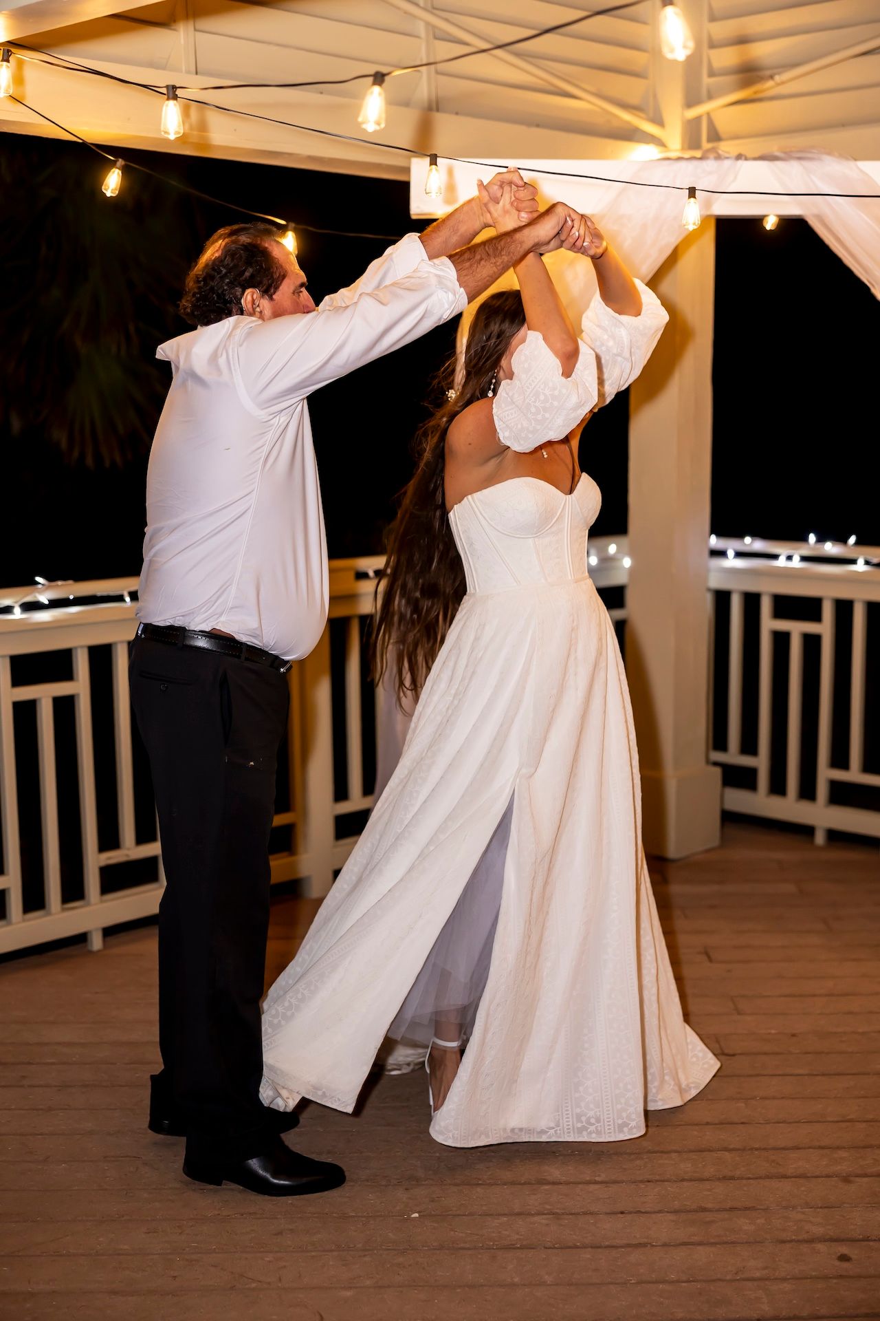 A couple in wedding attire dances joyfully on a wooden deck under string lights at an evening reception.