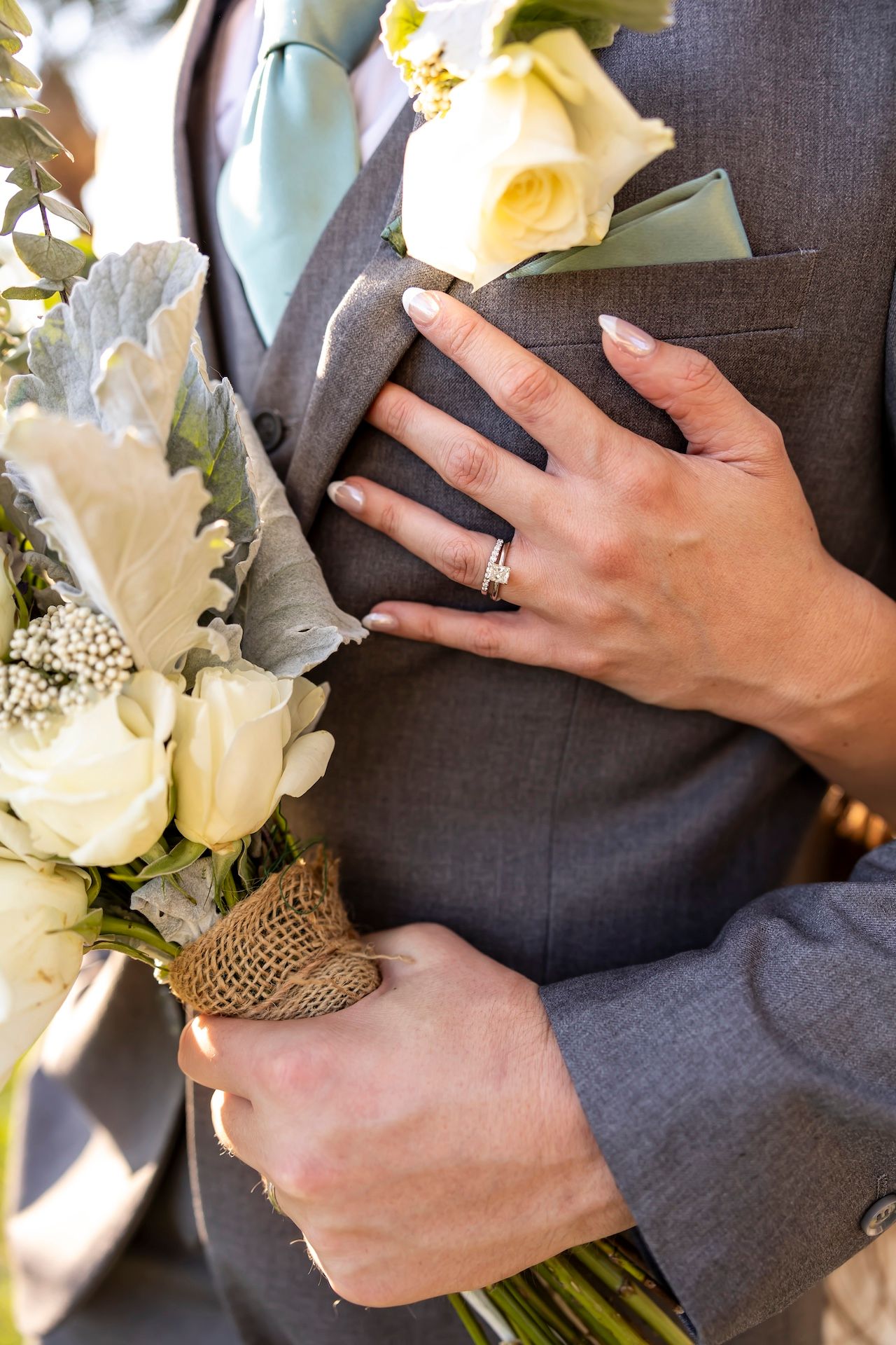 A hand wearing a wedding ring holds a bouquet wrapped in burlap against a grey suit with a mint tie.