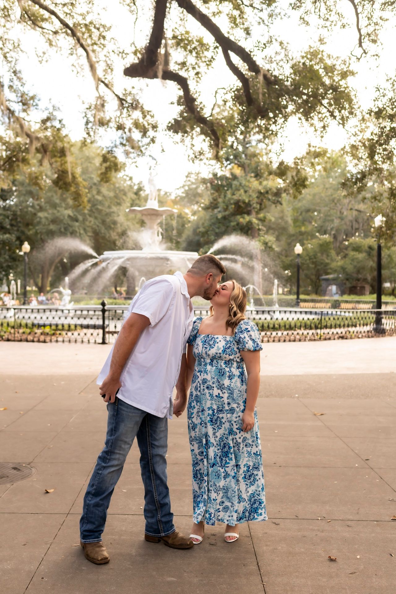 A couple shares a romantic moment in front of a fountain at a scenic park during golden hour.