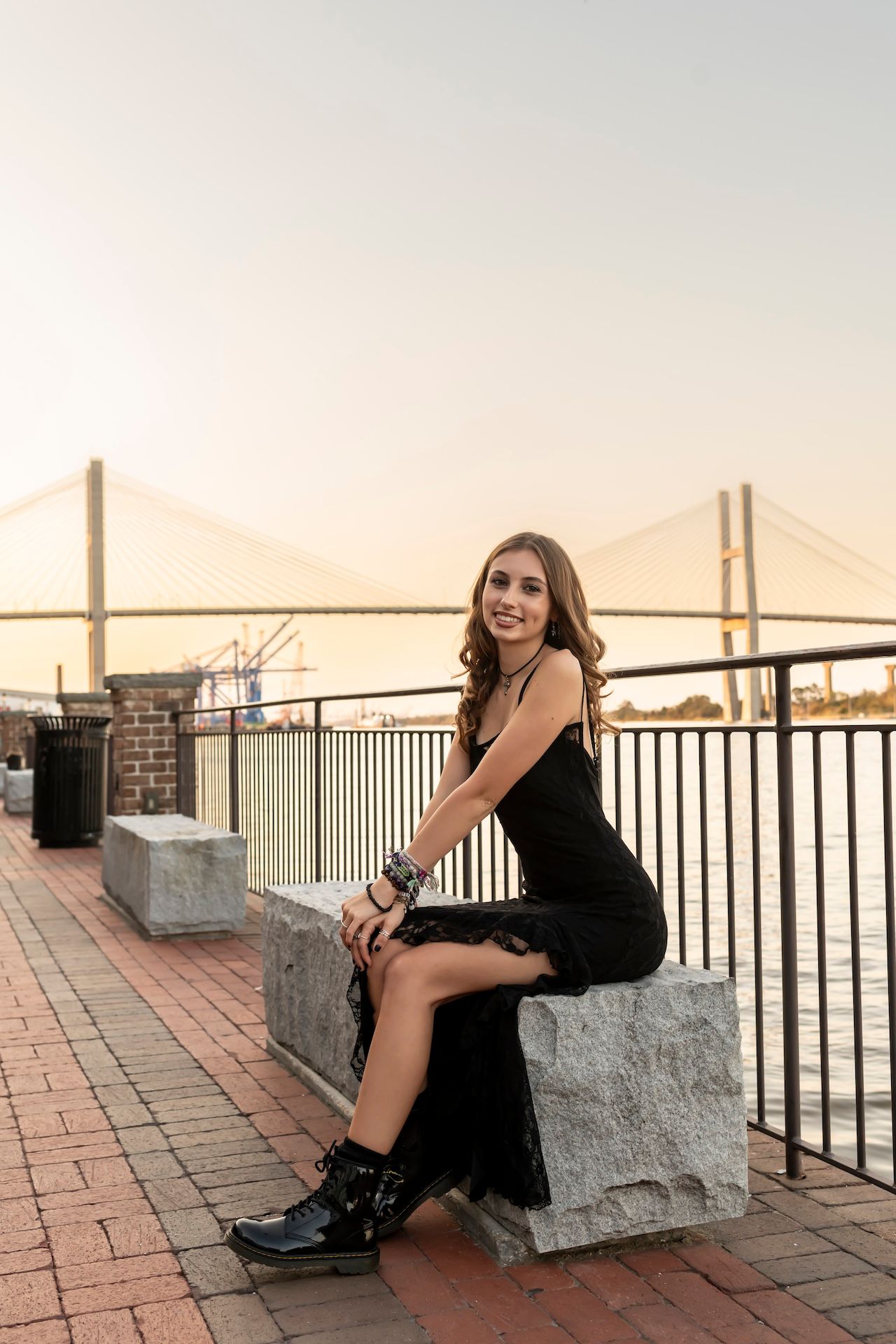 Person in a black dress sits on stone block along riverfront with suspension bridge in background at sunset.