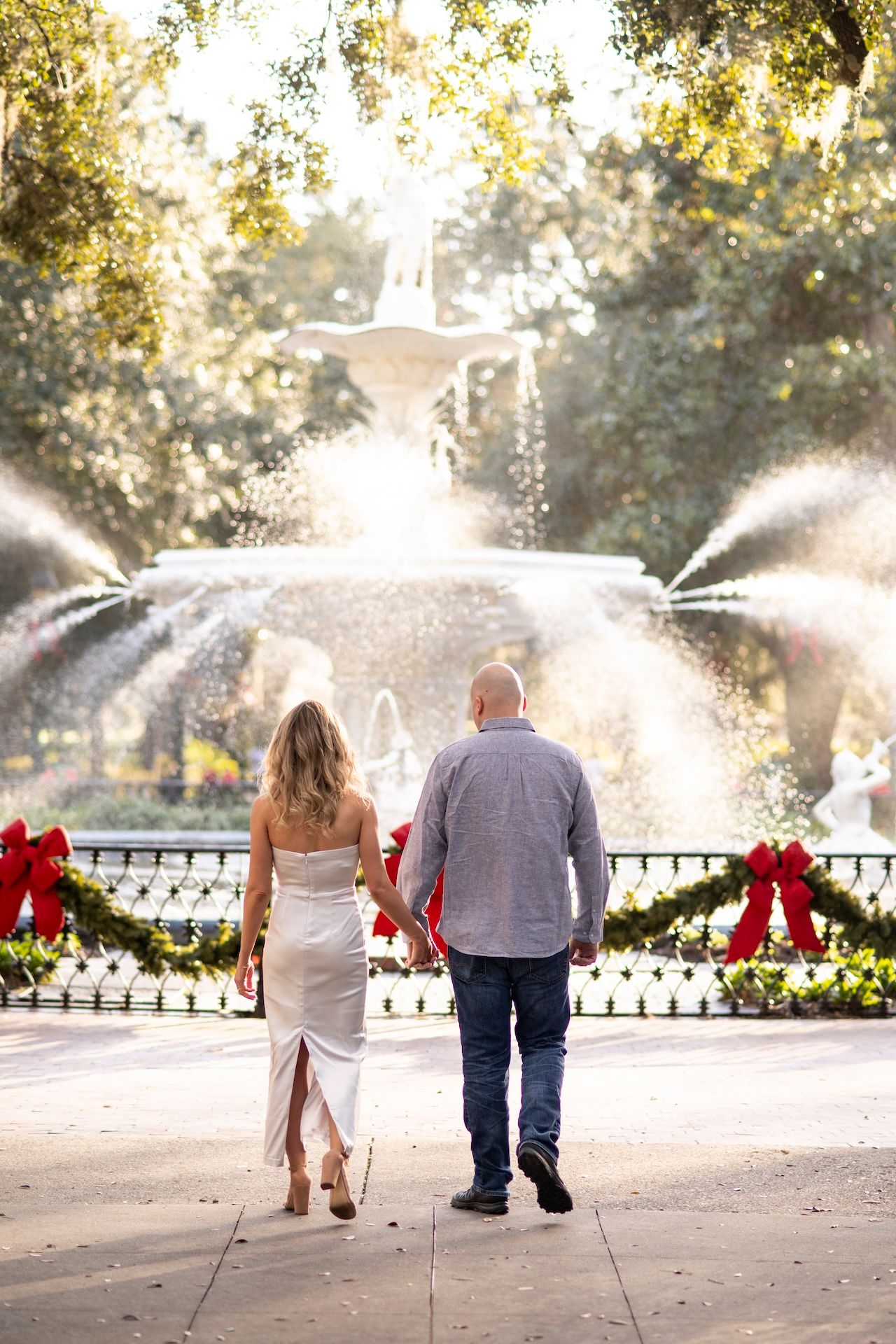 A couple walks together along a garden path with fountains and red holiday bows in the background on a sunny day.