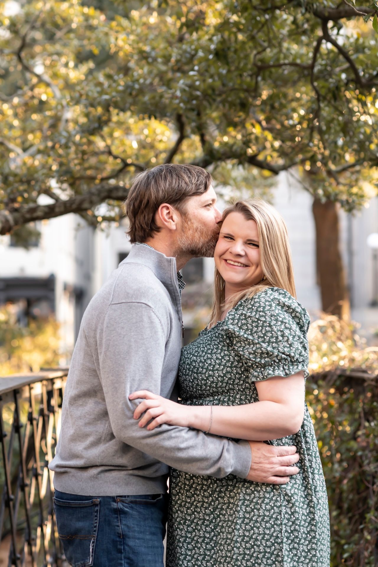 Couple embracing outdoors on a bridge with autumn foliage in background.