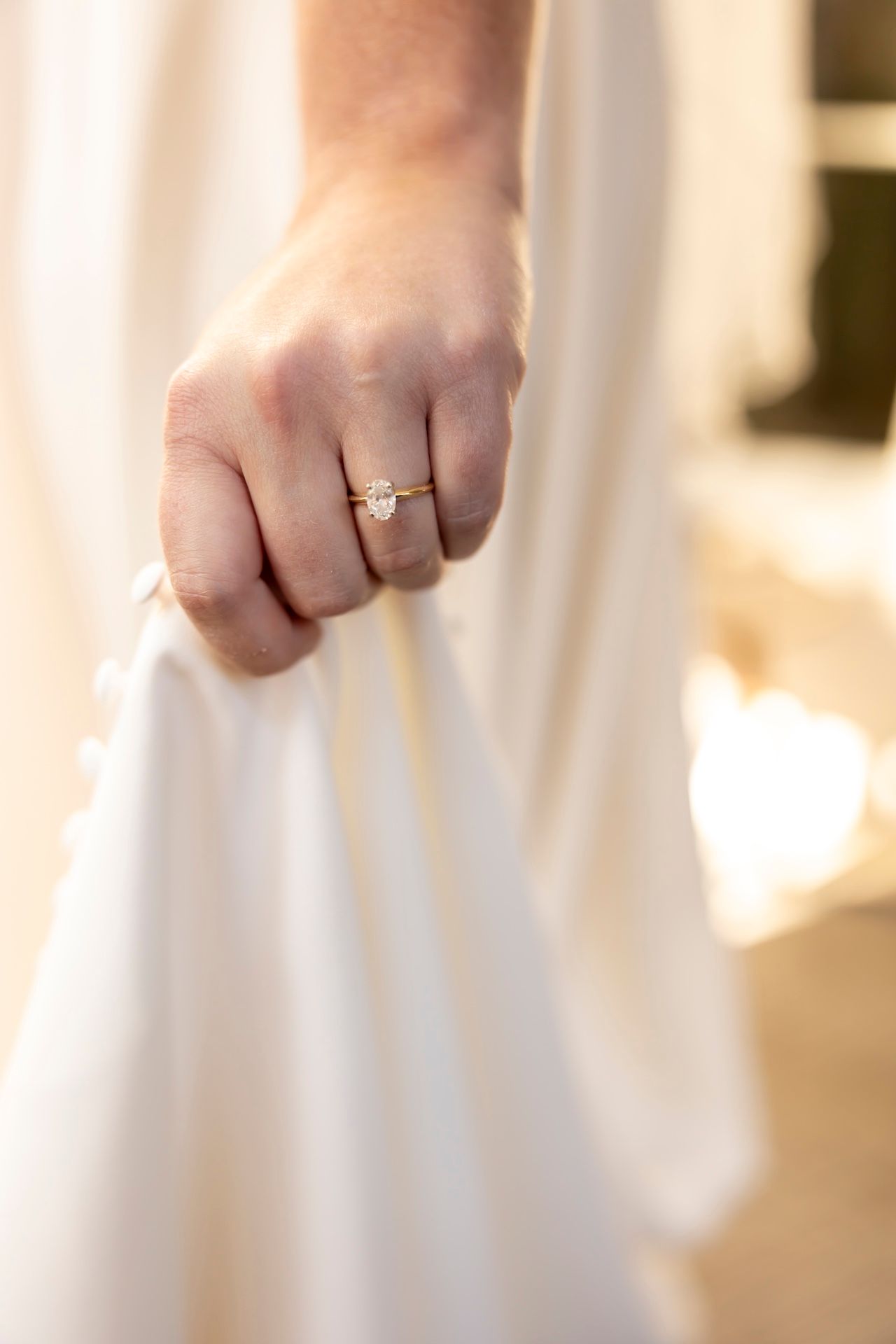 A delicate engagement ring sparkles on a hand against flowing white wedding dress fabric in soft lighting.