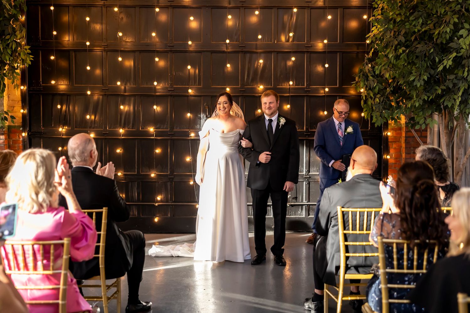 A romantic indoor wedding ceremony with string lights and greenery decorating a dark industrial style venue.