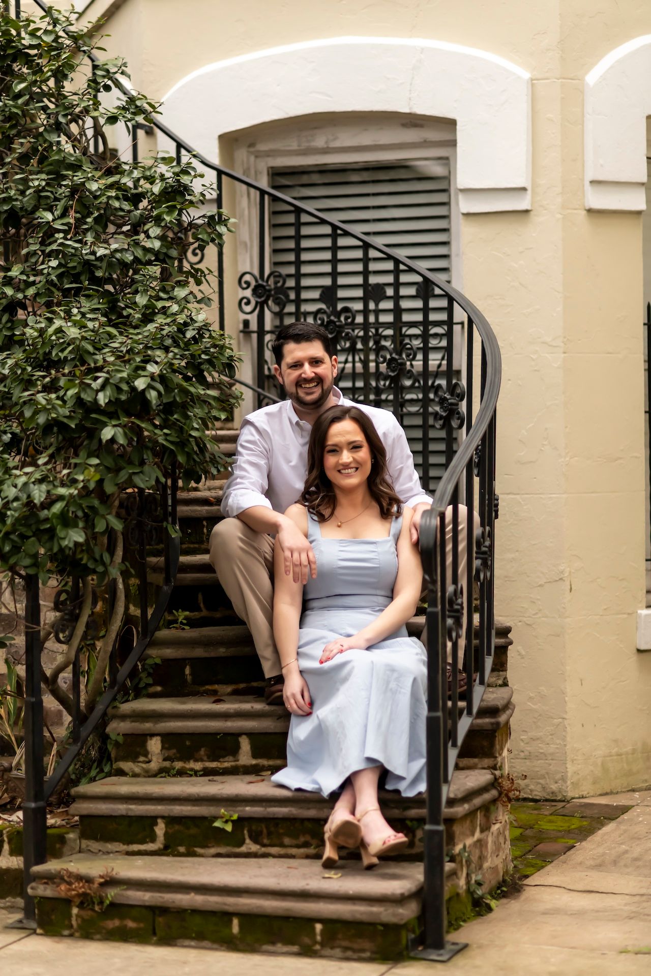 A couple sits together on an elegant curved outdoor staircase with wrought iron railings and greenery nearby.