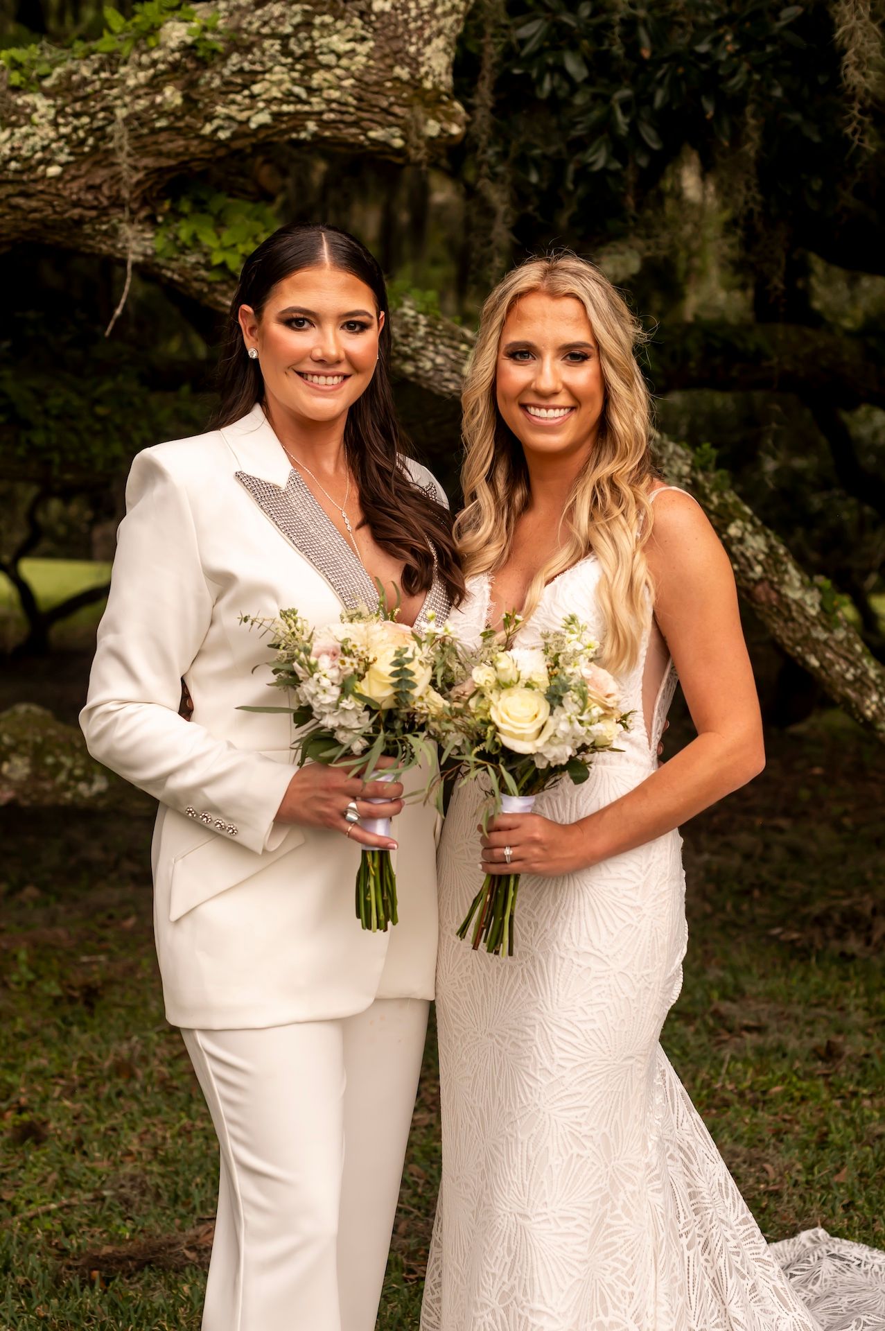 Two brides in white attire stand together holding matching bouquets of cream-colored flowers in an outdoor garden setting.
