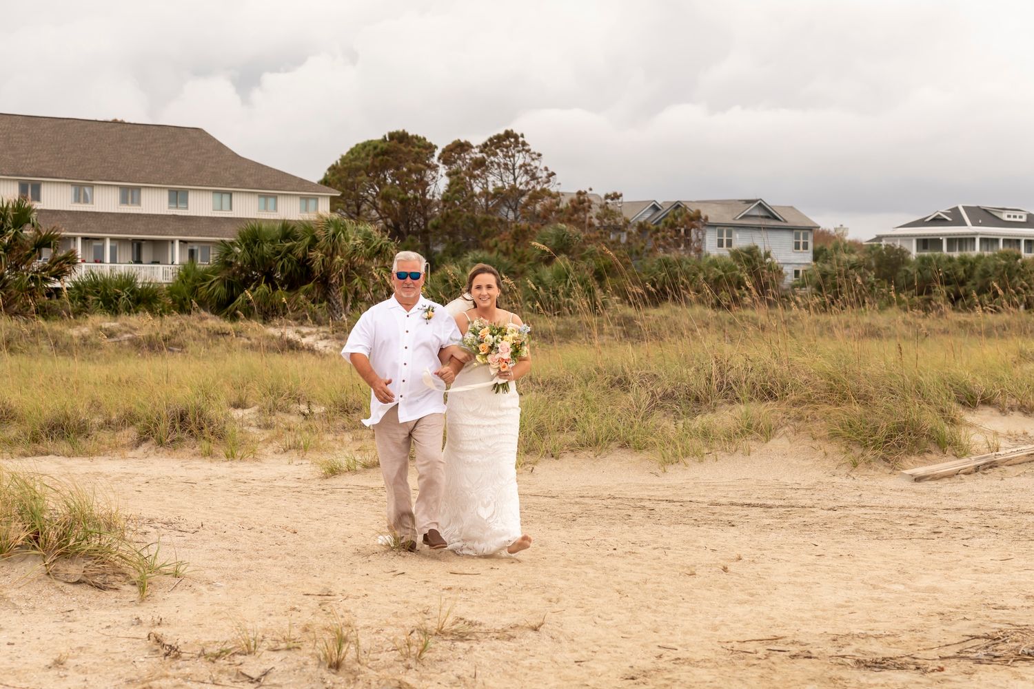 A couple walks hand in hand along a sandy beach path with coastal beach houses and dunes in the background.