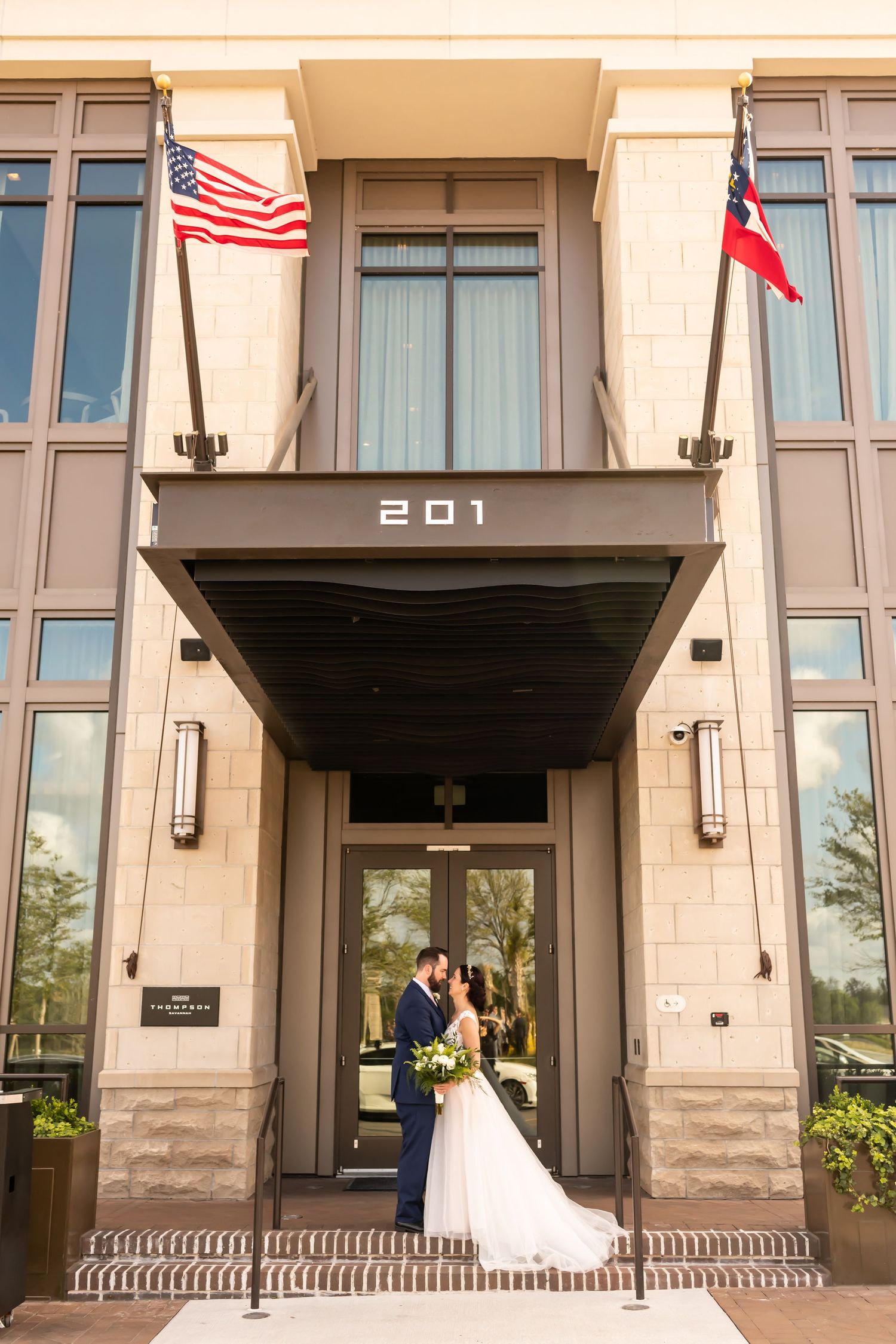 A couple in wedding attire stands beneath a beige building entrance with American and state flags displayed above.