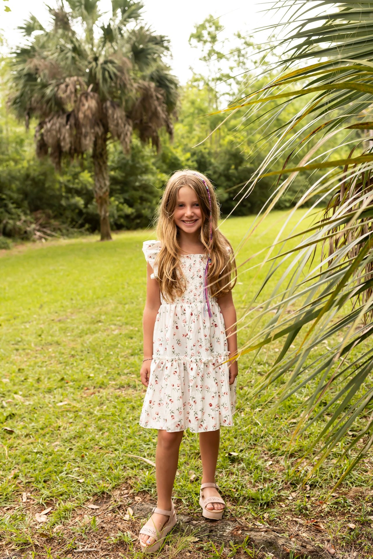 A young person in a white floral dress twirls joyfully in a grassy outdoor setting with palm trees.