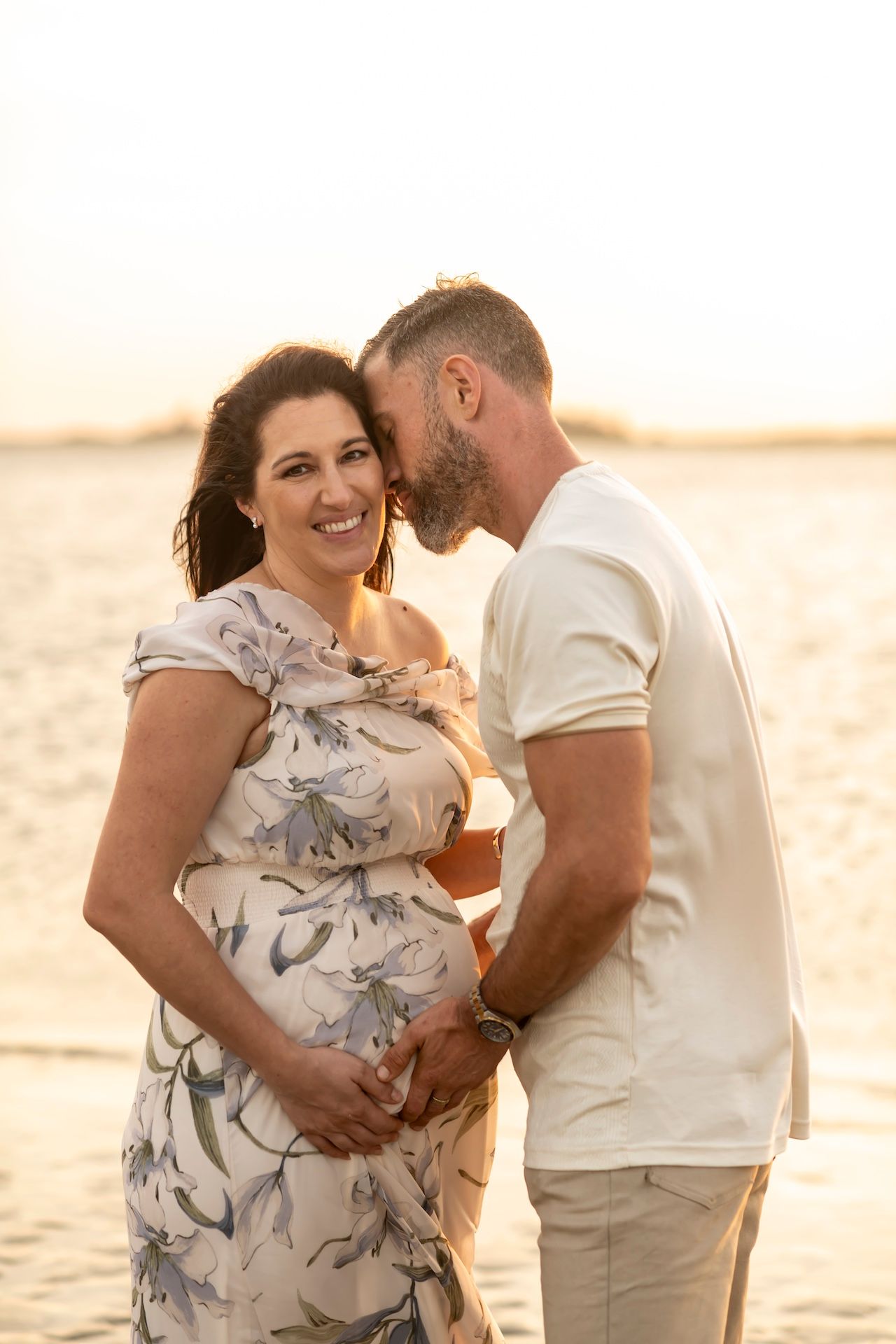 Romantic couple sharing an intimate moment on the beach at sunset while embracing her pregnant belly in matching light outfits.